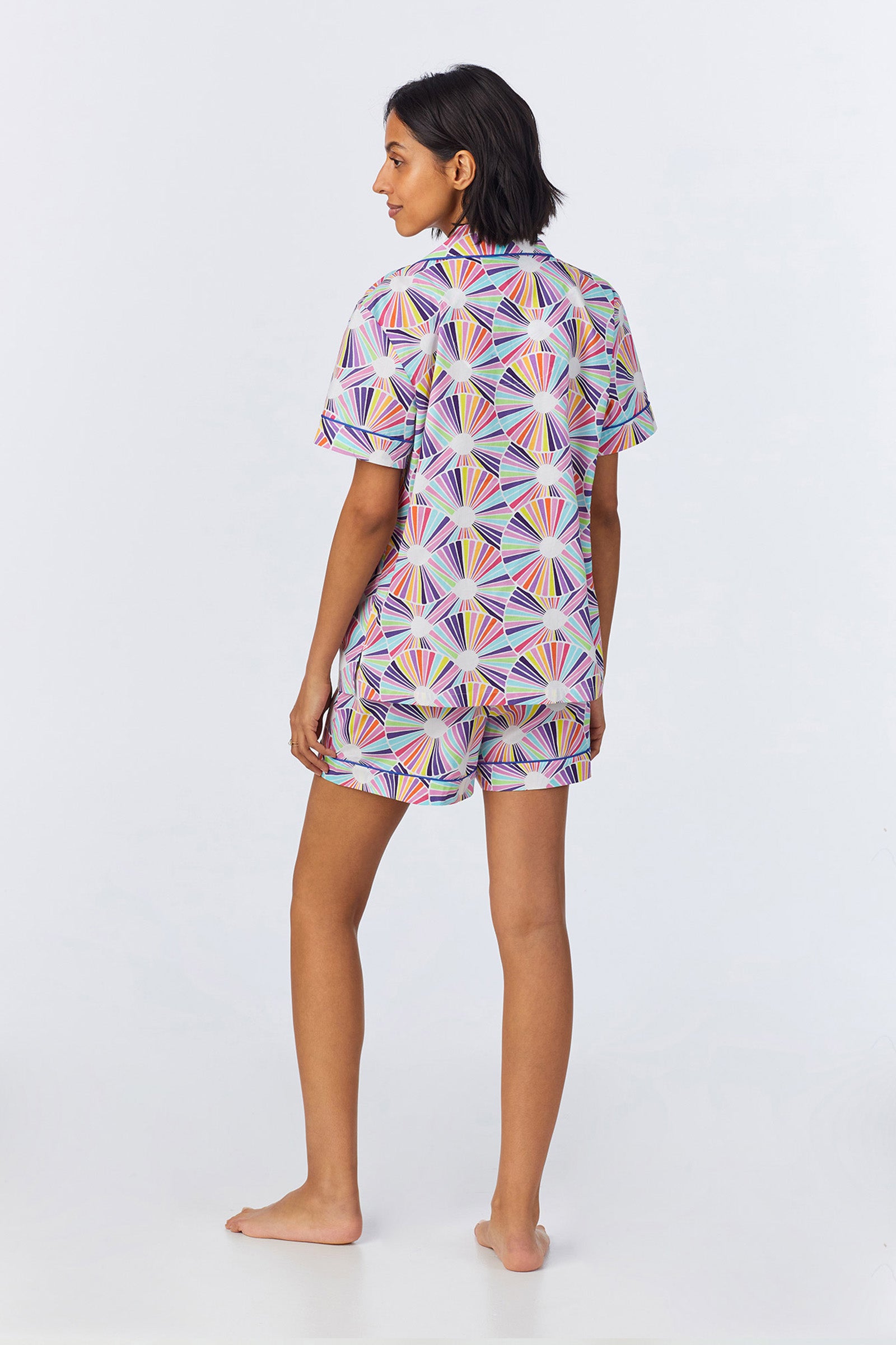 A person with short dark hair stands barefoot on a light background, facing away to show the back of the Shorty PJ Set - Fanfare Poplin, a matching short-sleeve shirt and shorts featuring a colorful geometric fan pattern.