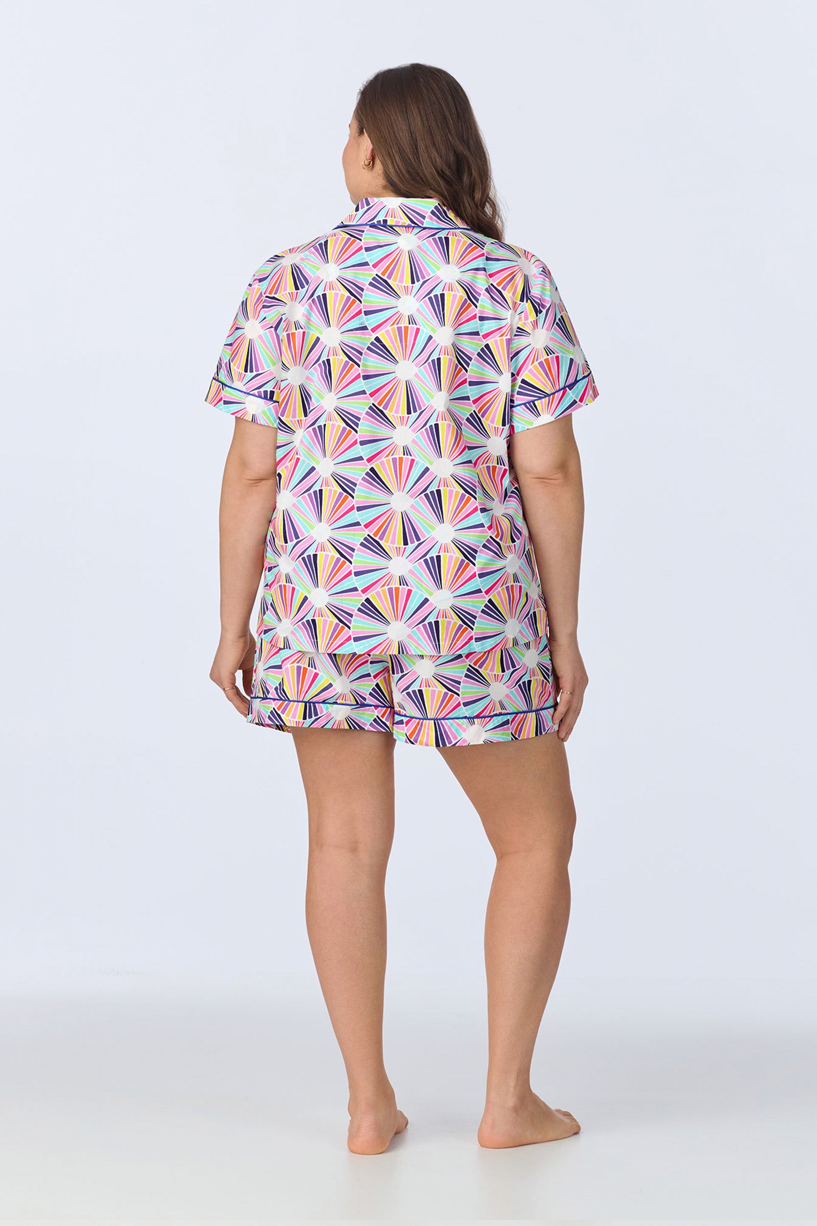 A woman stands barefoot, facing away, wearing the Shorty PJ Set - Fanfare Poplin with a colorful geometric fan pattern. The background is plain and light-colored.