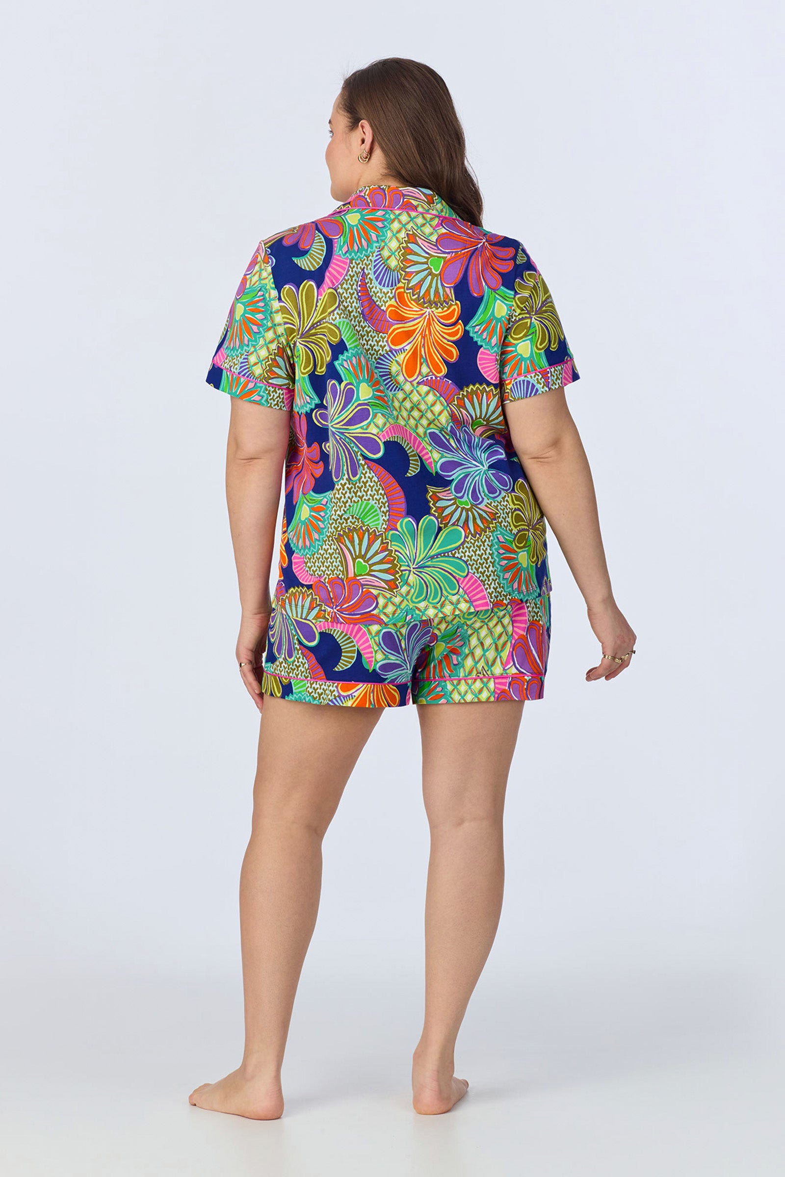 A woman faces away, barefoot, wearing the Shorty PJ Set - Zanzibar Stretch Jersey, featuring a bold green, orange, pink, and purple floral and leaf print on a navy background with short sleeves.