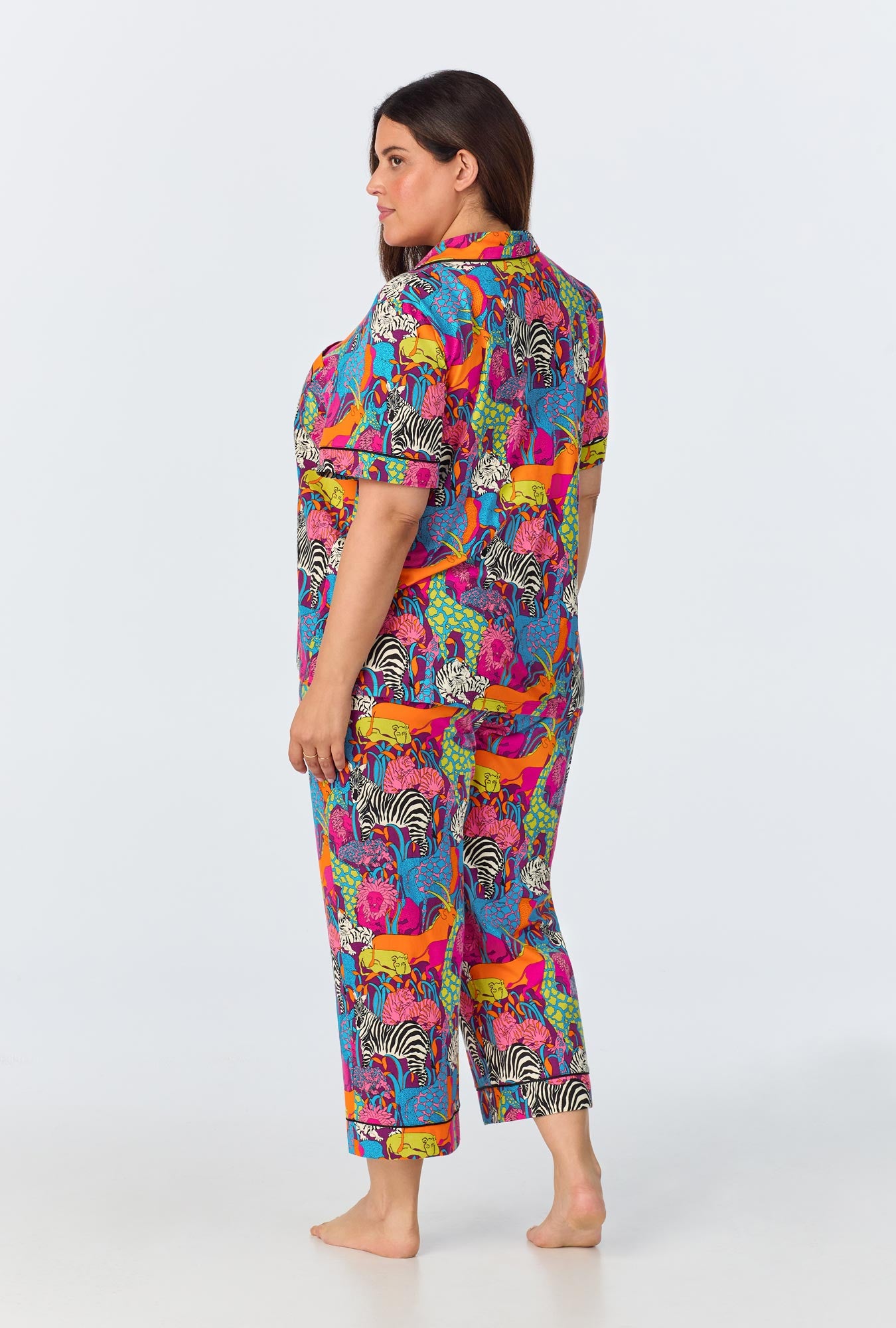 A woman in the Jungle Womens Short Sleeve Cropped Pant Jersey PJ Set stands barefoot, wearing colorful organic cotton pajamas with zebra, leopard, and jungle designs on a white background. She is turned slightly to the side, looking off-camera relaxed.