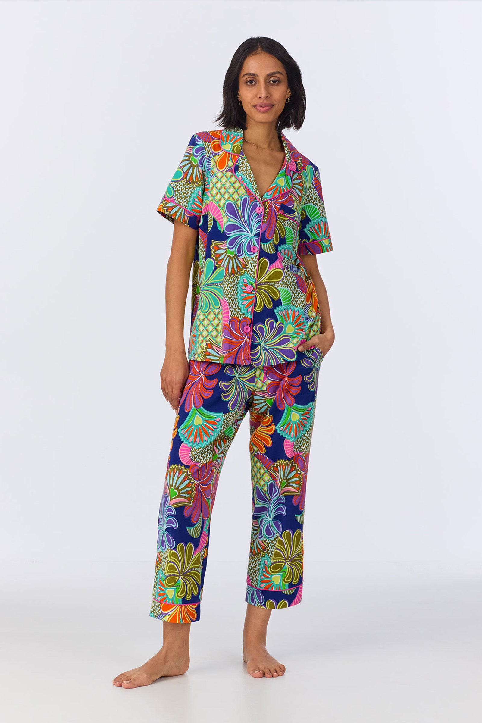 A woman stands barefoot against a plain background, wearing the Crop Classic PJ Set - Zanzibar Stretch Jersey, featuring a bold floral pattern in orange, green, purple, blue, and pink.