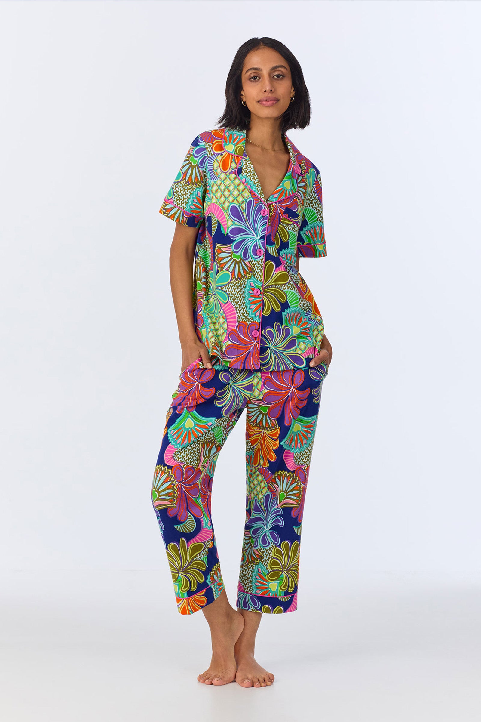 A woman stands barefoot against a plain background, wearing the Crop Classic PJ Set - Zanzibar Stretch Jersey in Multi, featuring a bold and colorful floral and leaf pattern.