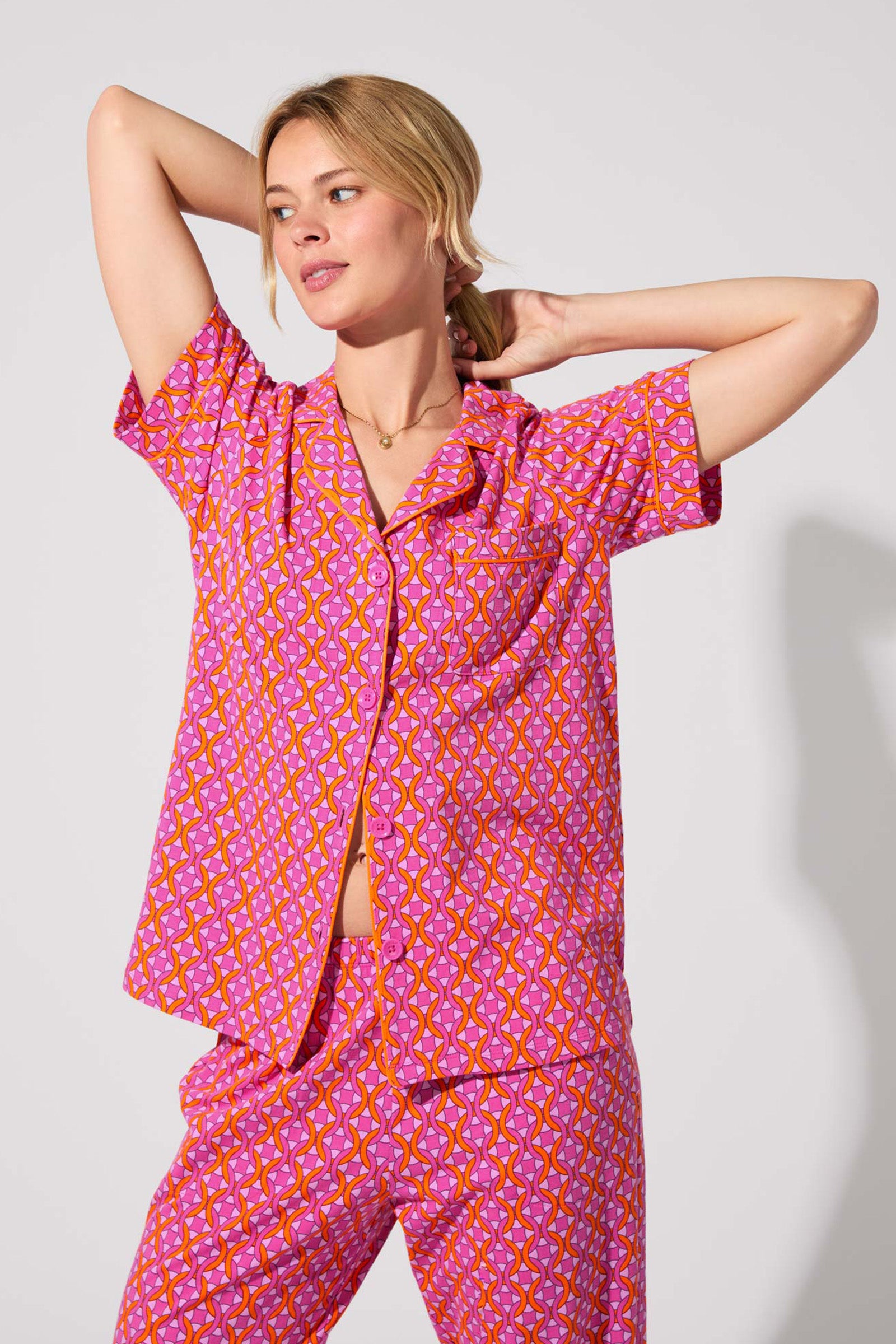 A person wears the Crop Classic PJ Set - Chain Geo, a pink and orange patterned short-sleeve pajama set, standing with arms raised and looking to the side against a plain light background.