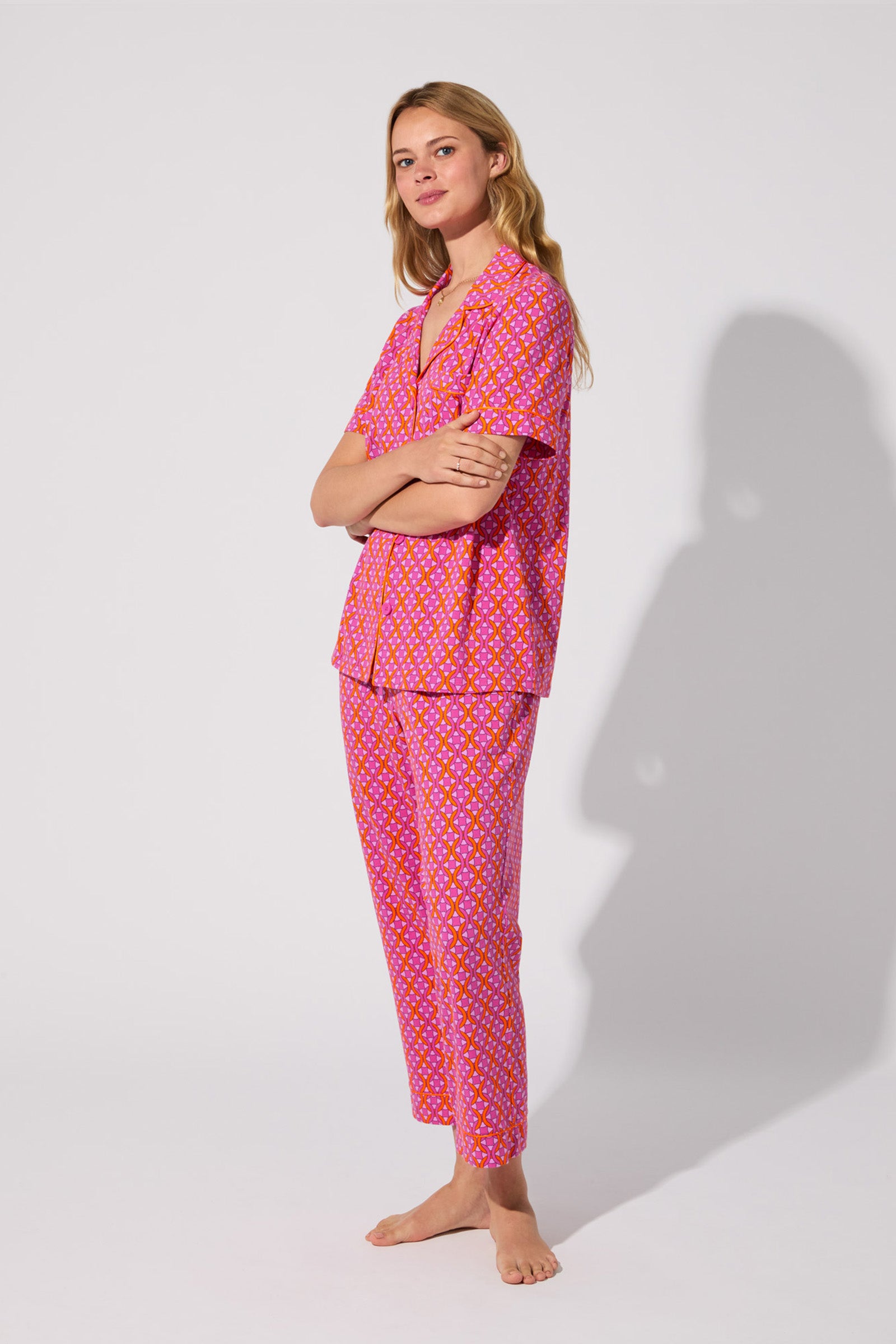 A woman stands barefoot against a plain background, smiling softly with her arms crossed. She wears the Crop Classic PJ Set - Chain Geo, a matching pink pajama set featuring a yellow and white polka dot pattern. Her long hair is loose.