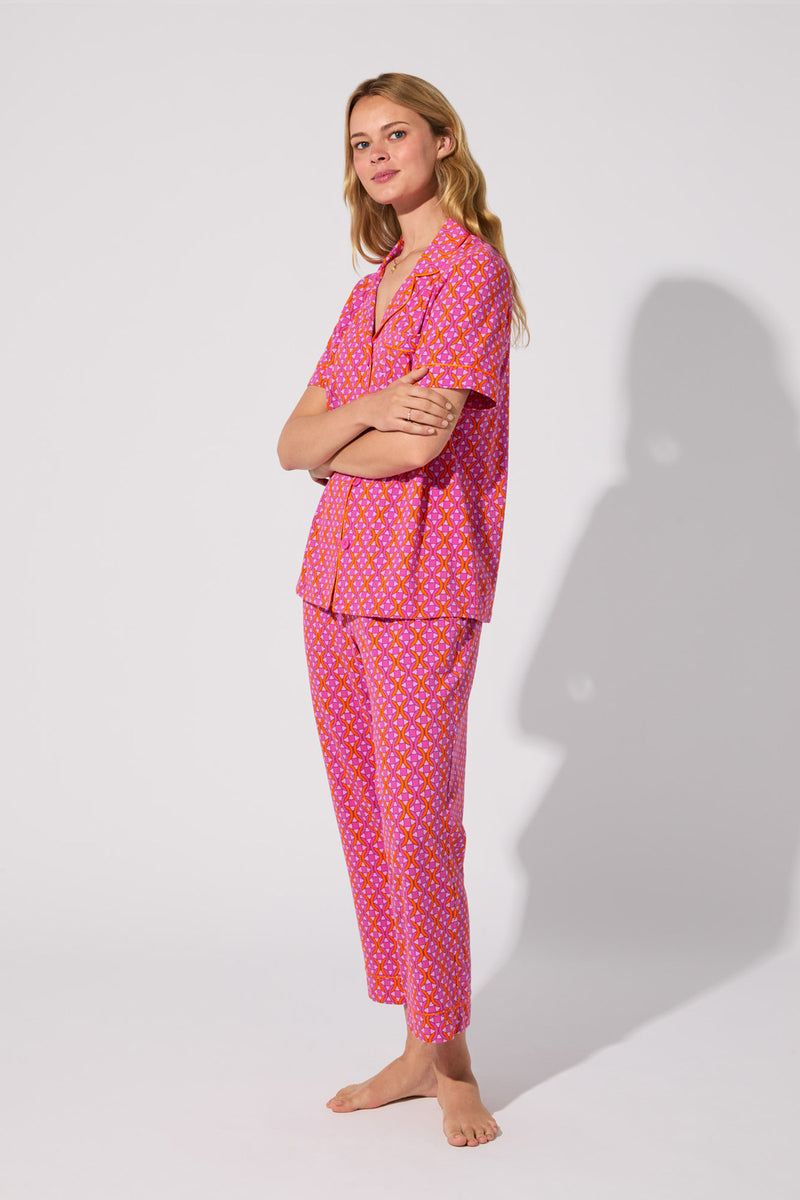 A woman stands barefoot against a plain background, smiling softly with her arms crossed. She wears the Crop Classic PJ Set - Chain Geo, a matching pink pajama set featuring a yellow and white polka dot pattern. Her long hair is loose.