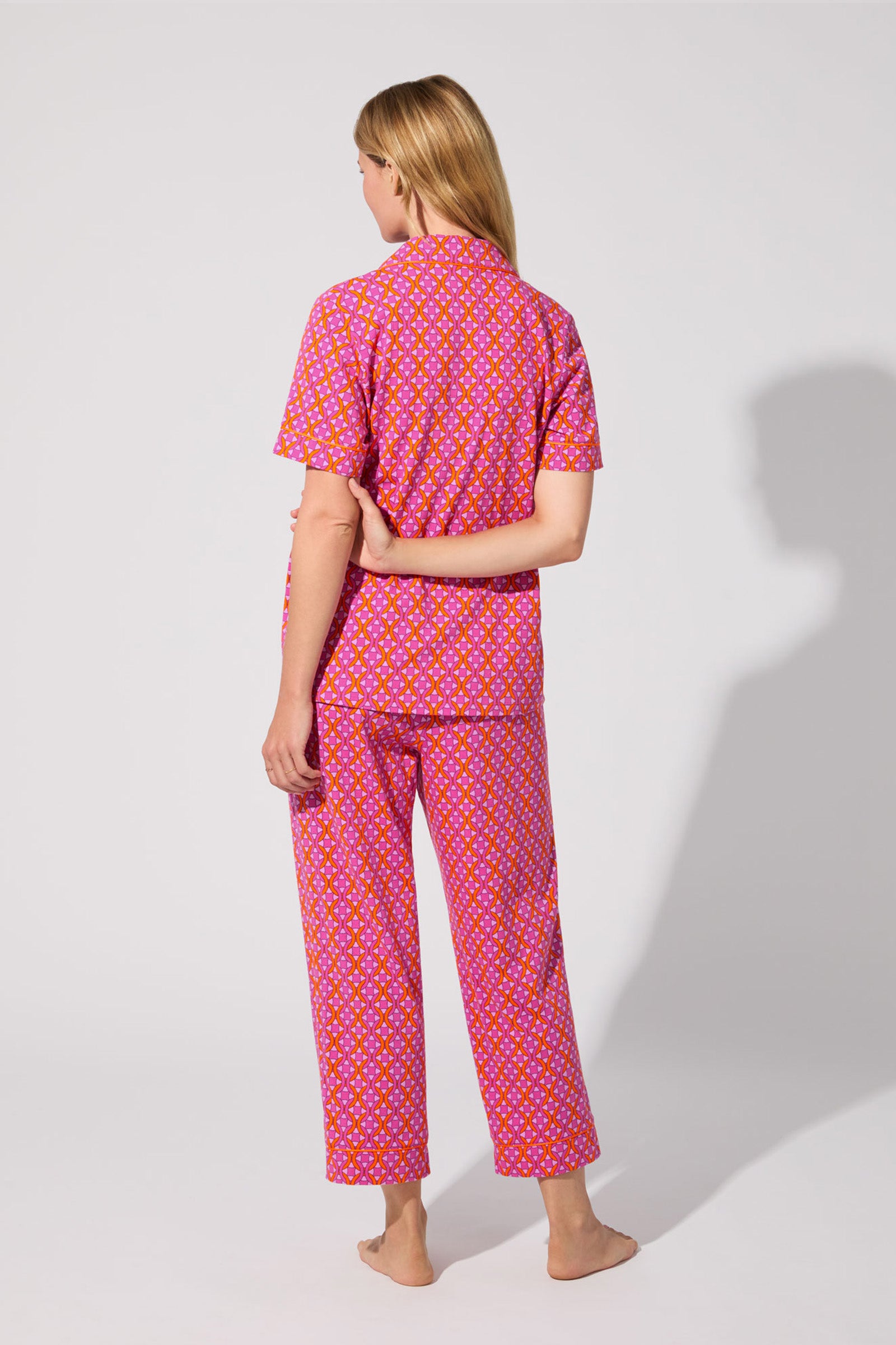 A person with long blonde hair stands barefoot facing away, wearing the Crop Classic PJ Set - Chain Geo, a short-sleeved, button-up pajama set. Shadows fall across the light gray floor and wall.