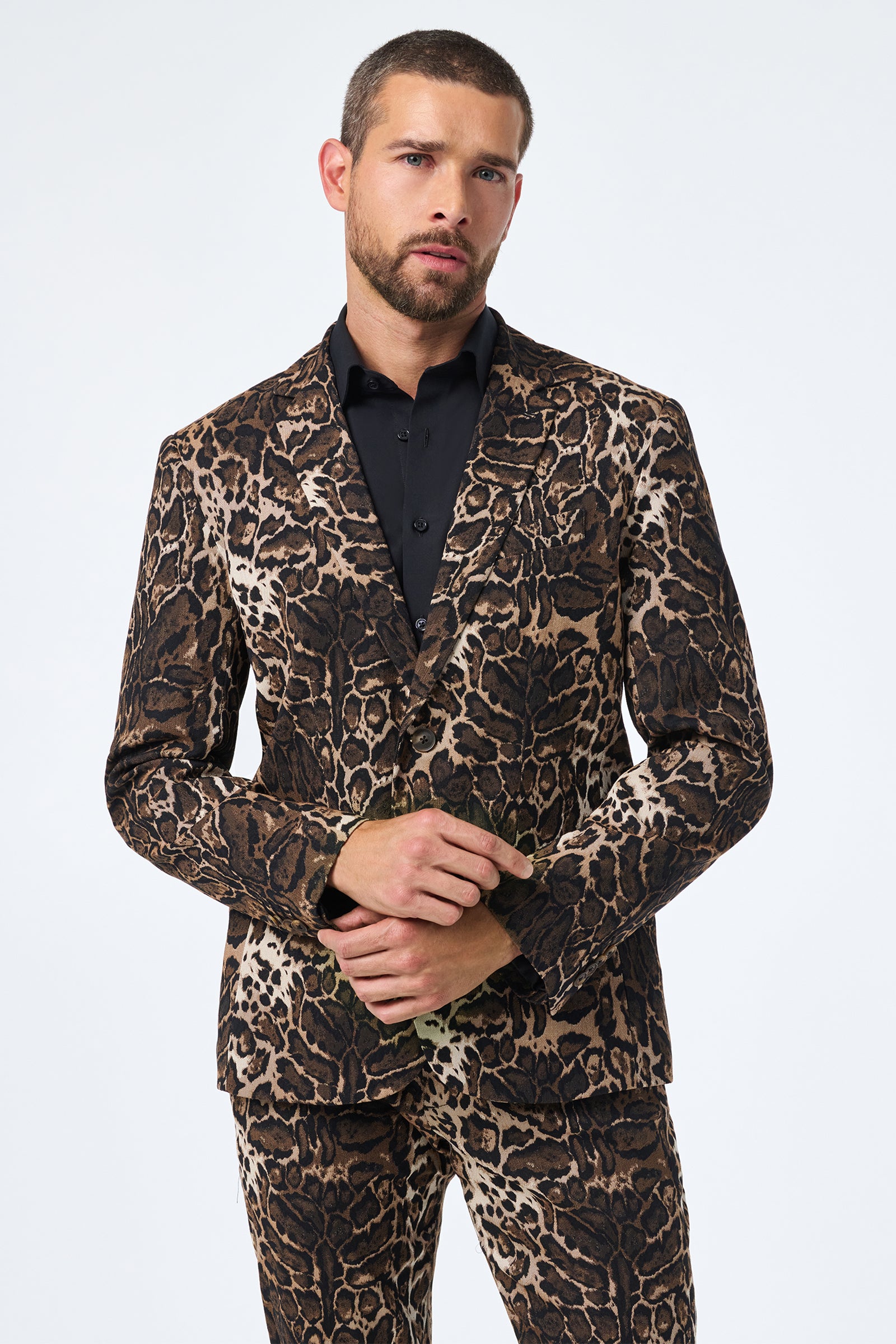 A man with short hair and a beard poses against a plain light background, hands lightly clasped, wearing the Thurston Blazer in Lompoul Leopard over a black dress shirt.