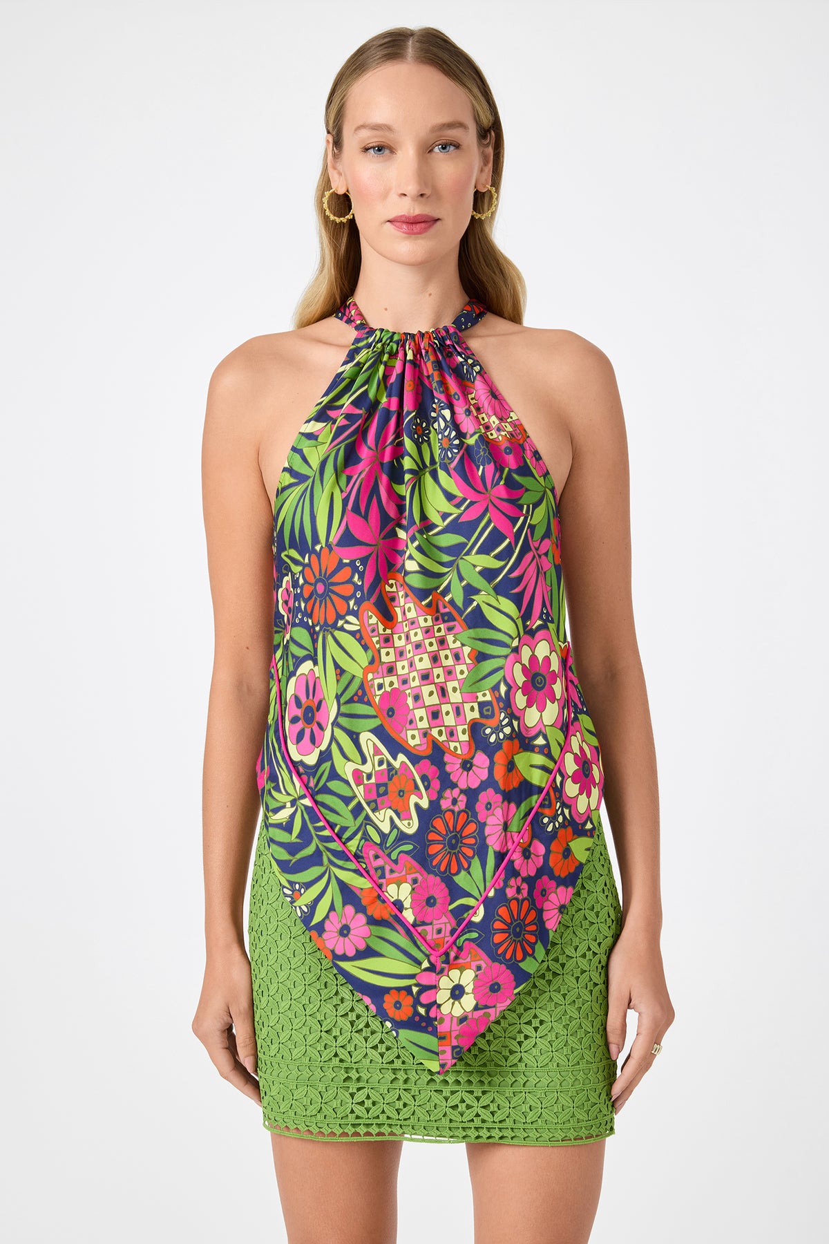 A woman wearing the Lavani 2 Halter Top - Fumi Floral and a fitted green lace skirt stands against a white background. She has long, wavy blonde hair and wears gold hoop earrings.