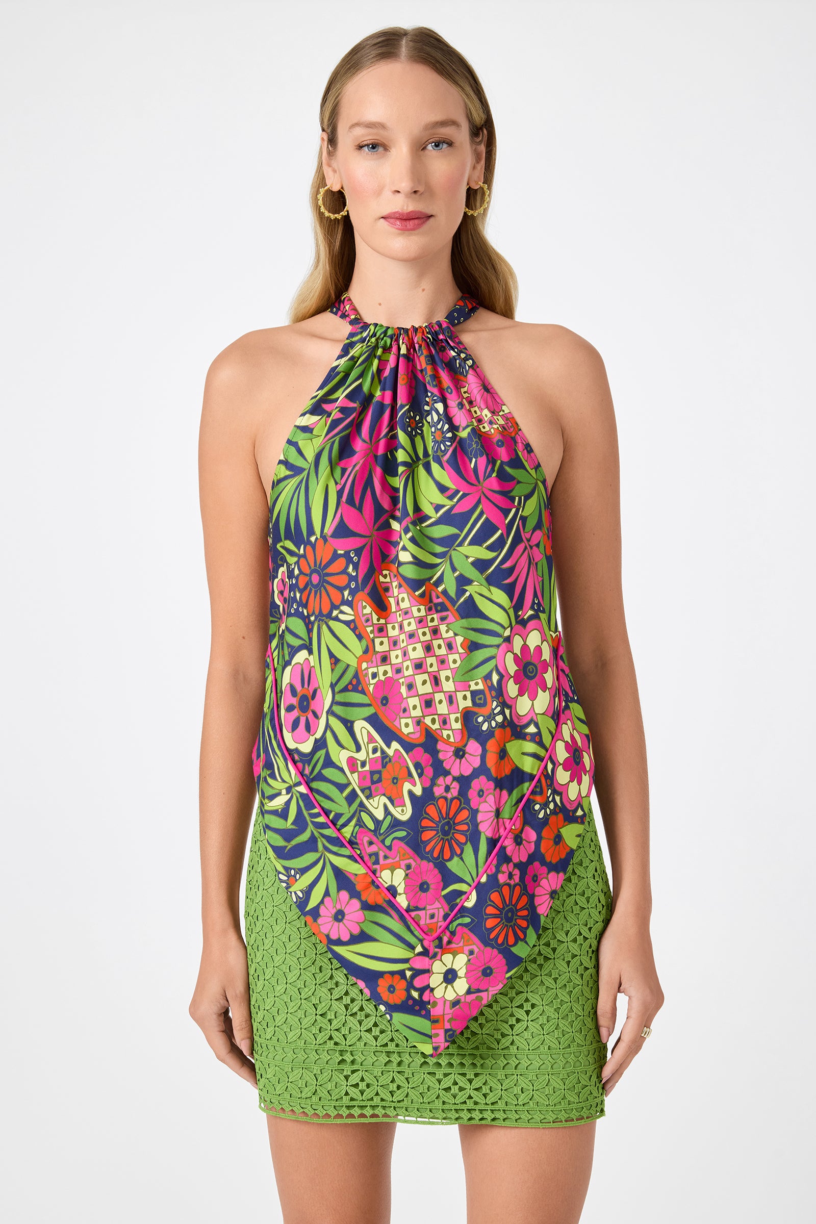 A woman wearing the Lavani 2 Halter Top - Fumi Floral and a fitted green lace skirt stands against a white background. She has long, wavy blonde hair and wears gold hoop earrings.