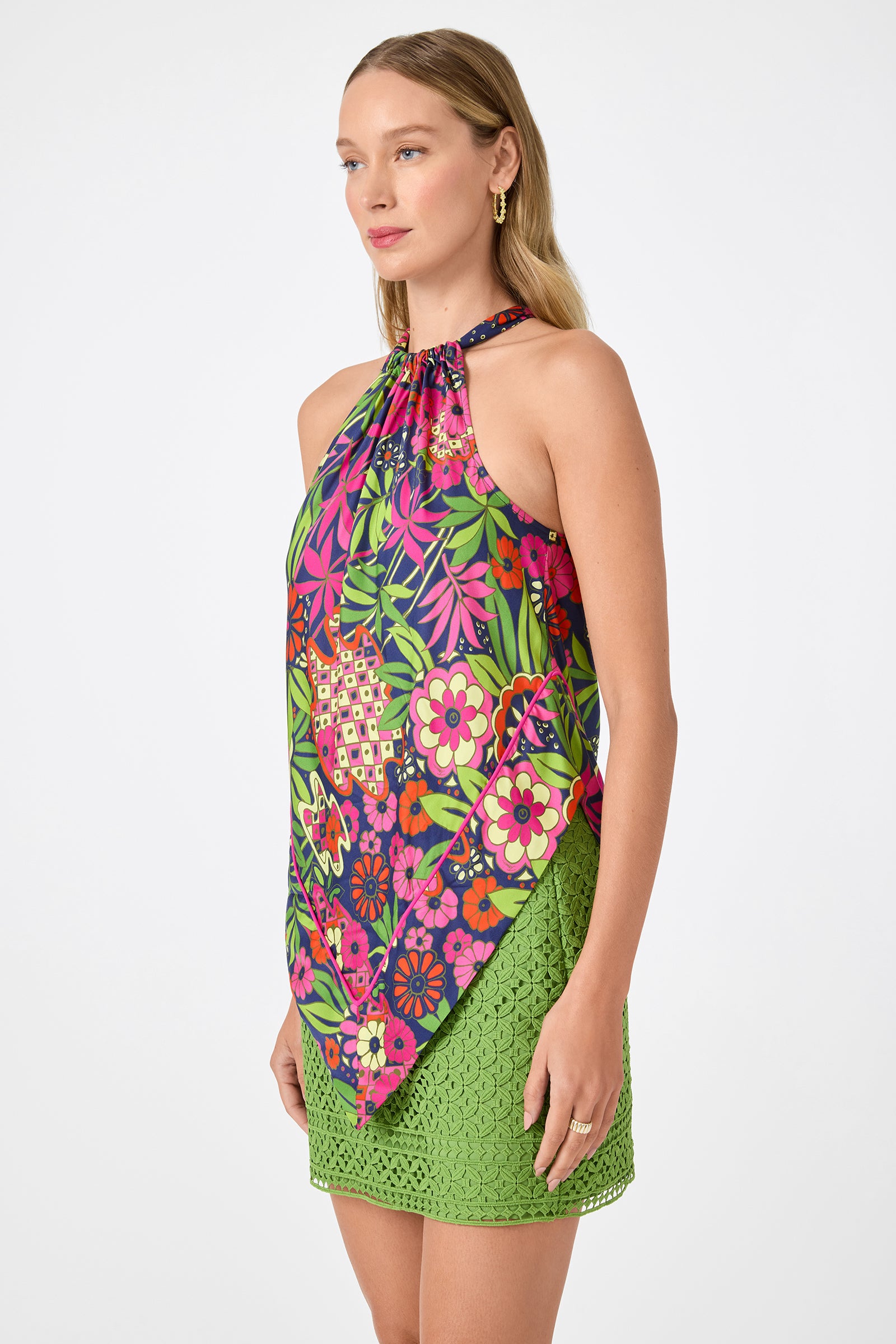 A woman stands against a plain background wearing the Lavani 2 Halter Top in Fumi Floral, paired with a green lace mini skirt. She has long, straight blonde hair and gold hoop earrings.