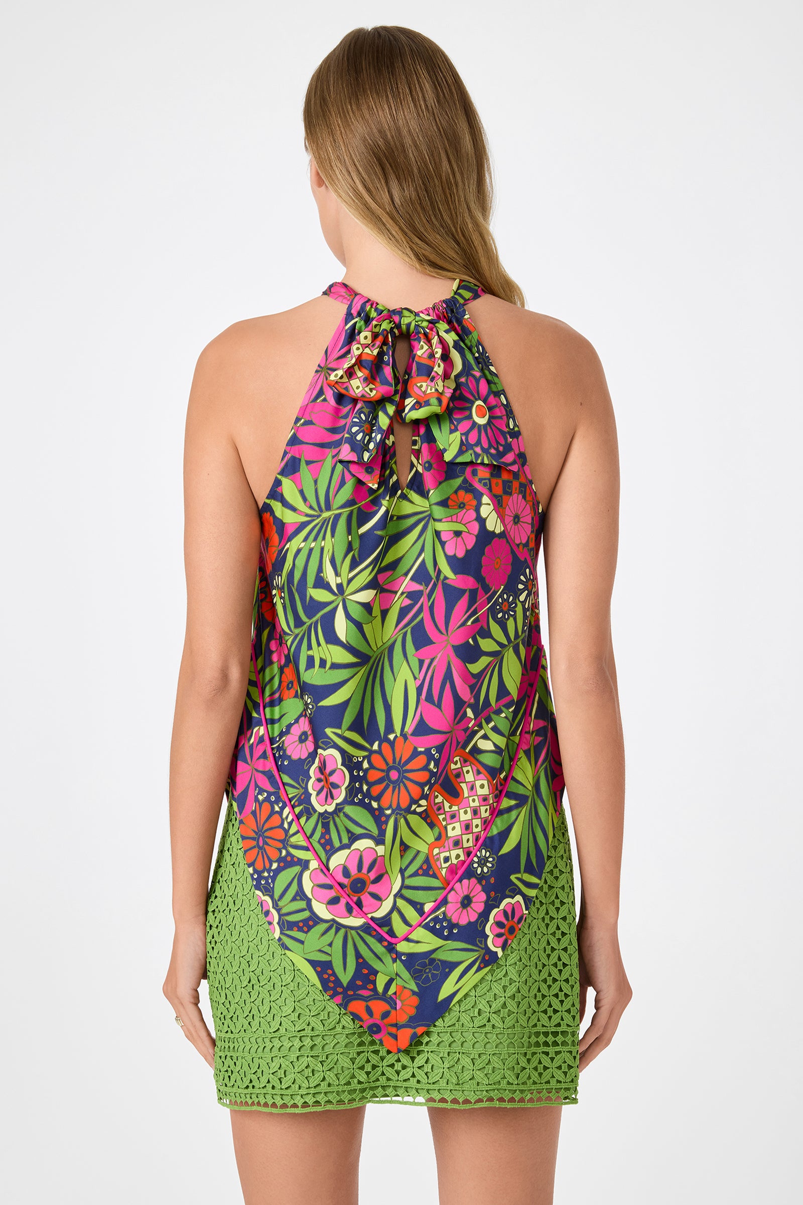 A woman with long light brown hair is seen from the back wearing the Lavani 2 Halter Top - Fumi Floral, featuring bright pink, green, and orange patterns on a dark blue base, paired with a green crochet skirt.
