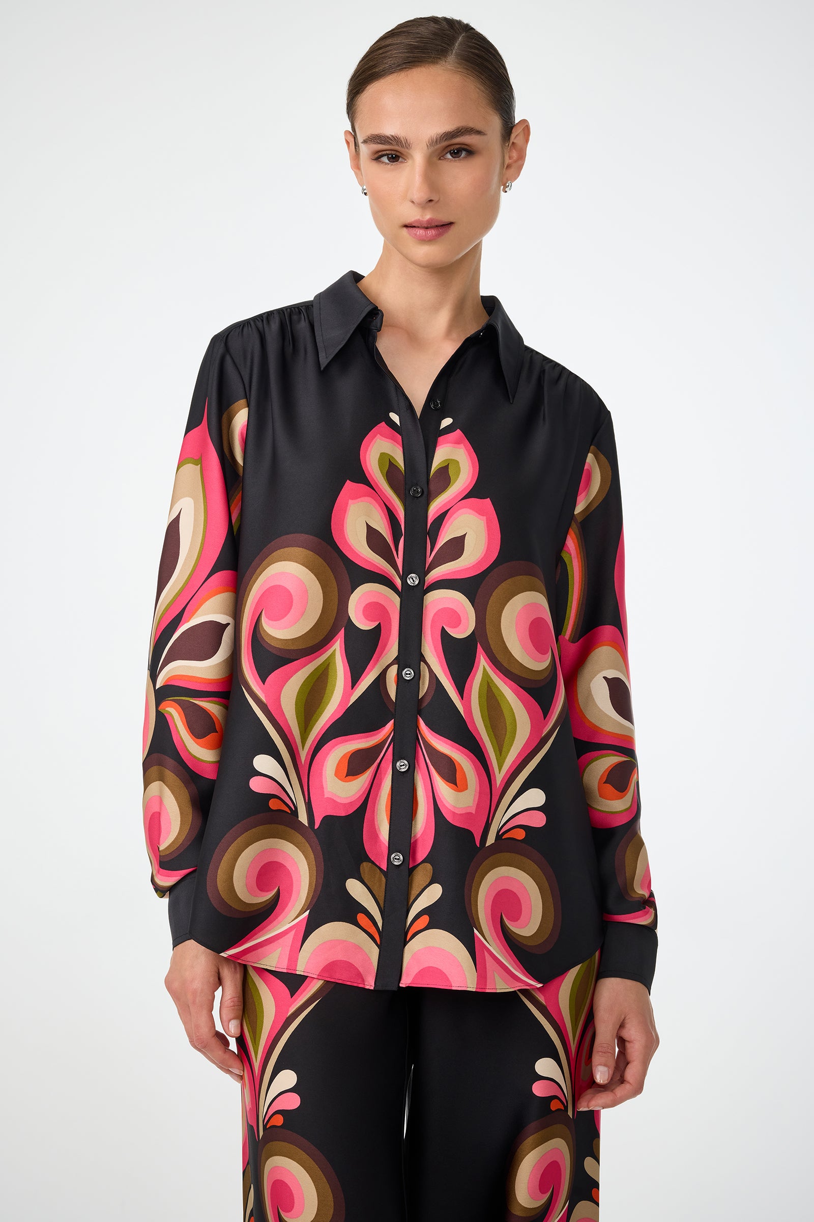 A woman with brown hair pulled back wears the Cassavetes Border Shirt in Sanctuary, featuring a long-sleeve button-up style with a bold, colorful abstract pattern on a black background.