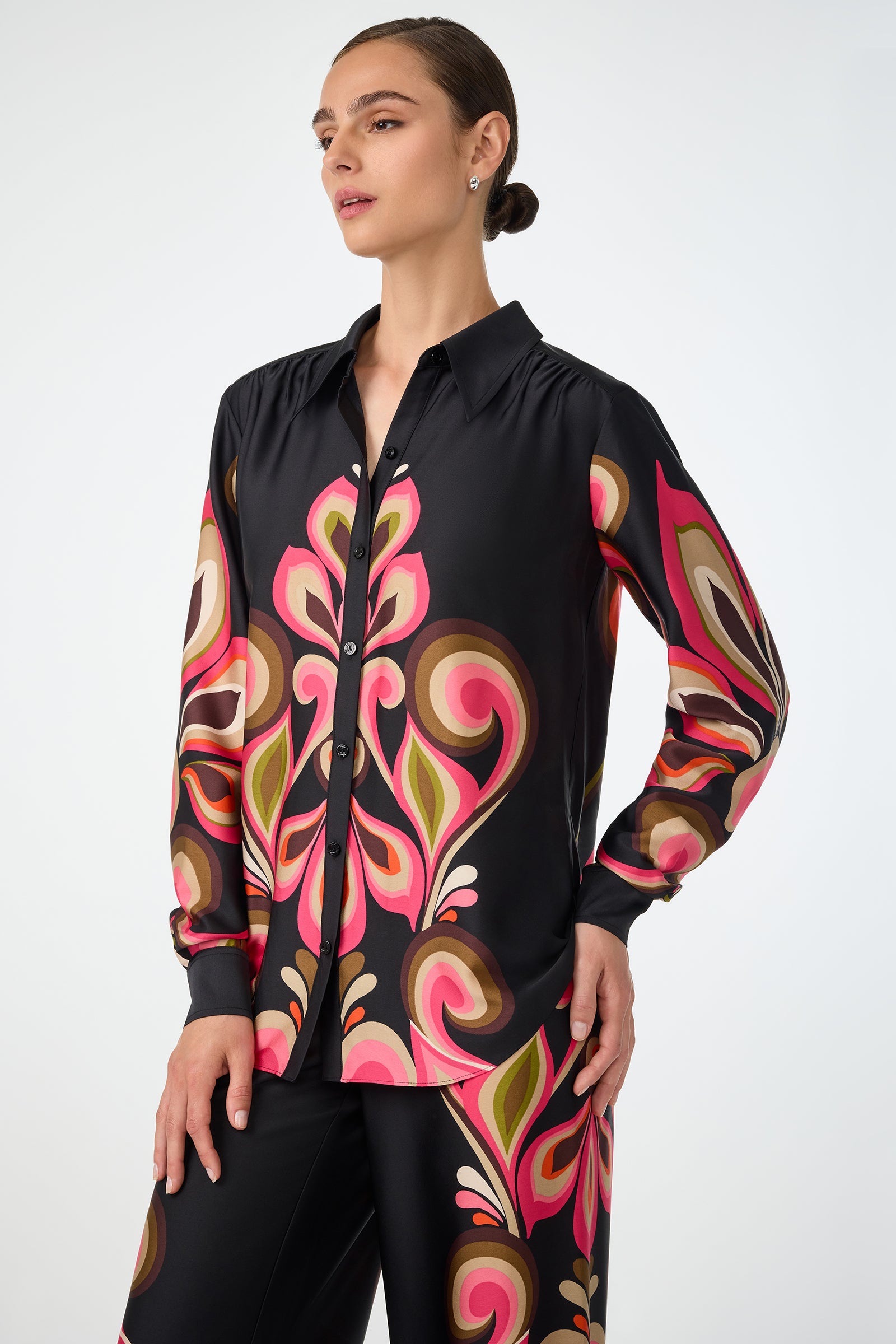 A woman wears the Cassavetes Border Shirt - Sanctuary, a black button-up with bold, swirling patterns in pink, red, green, and beige, paired with matching pants. She stands with one hand on her hip, looking to the side.