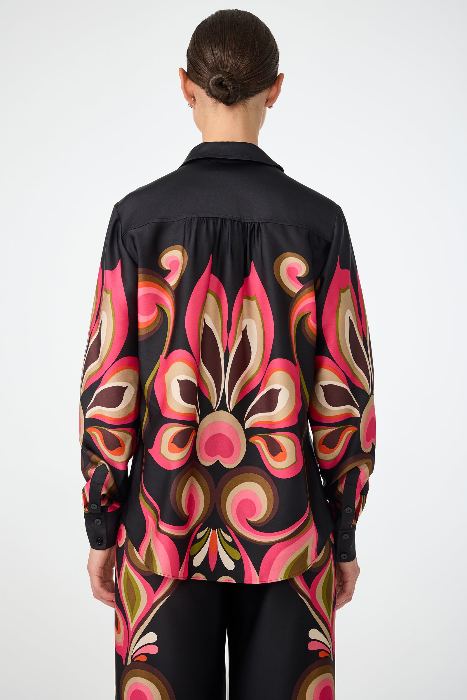 A person with dark hair in a low bun is shown from the back wearing the Cassavetes Border Shirt - Sanctuary, a black shirt and pants set with a bold, retro-inspired pink, brown, cream, and olive pattern against a plain background.