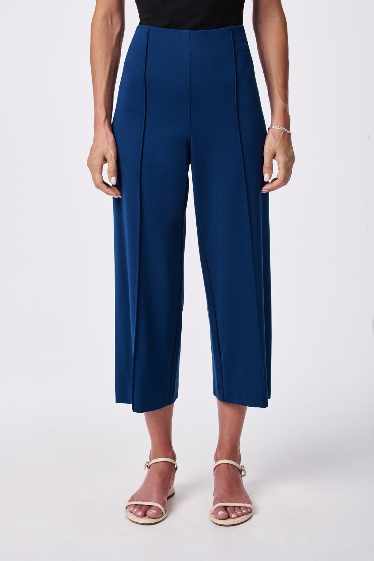 A person wearing the Santo Pant, a high-waisted, wide-leg cropped blue pant, paired with a black top, white strappy sandals, and a silver bracelet, stands against a plain white background.