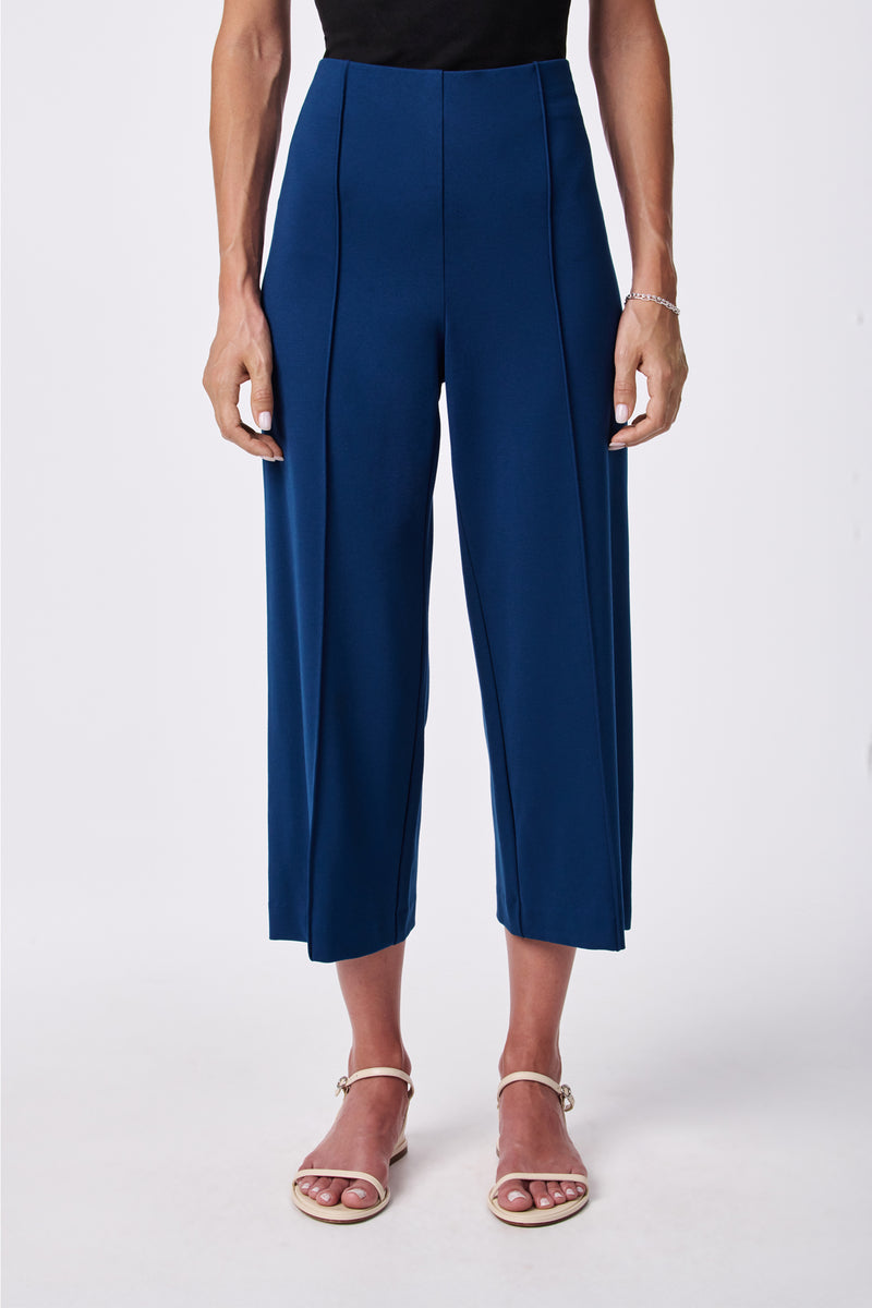 A person wearing the Santo Pant, a high-waisted, wide-leg cropped blue pant, paired with a black top, white strappy sandals, and a silver bracelet, stands against a plain white background.