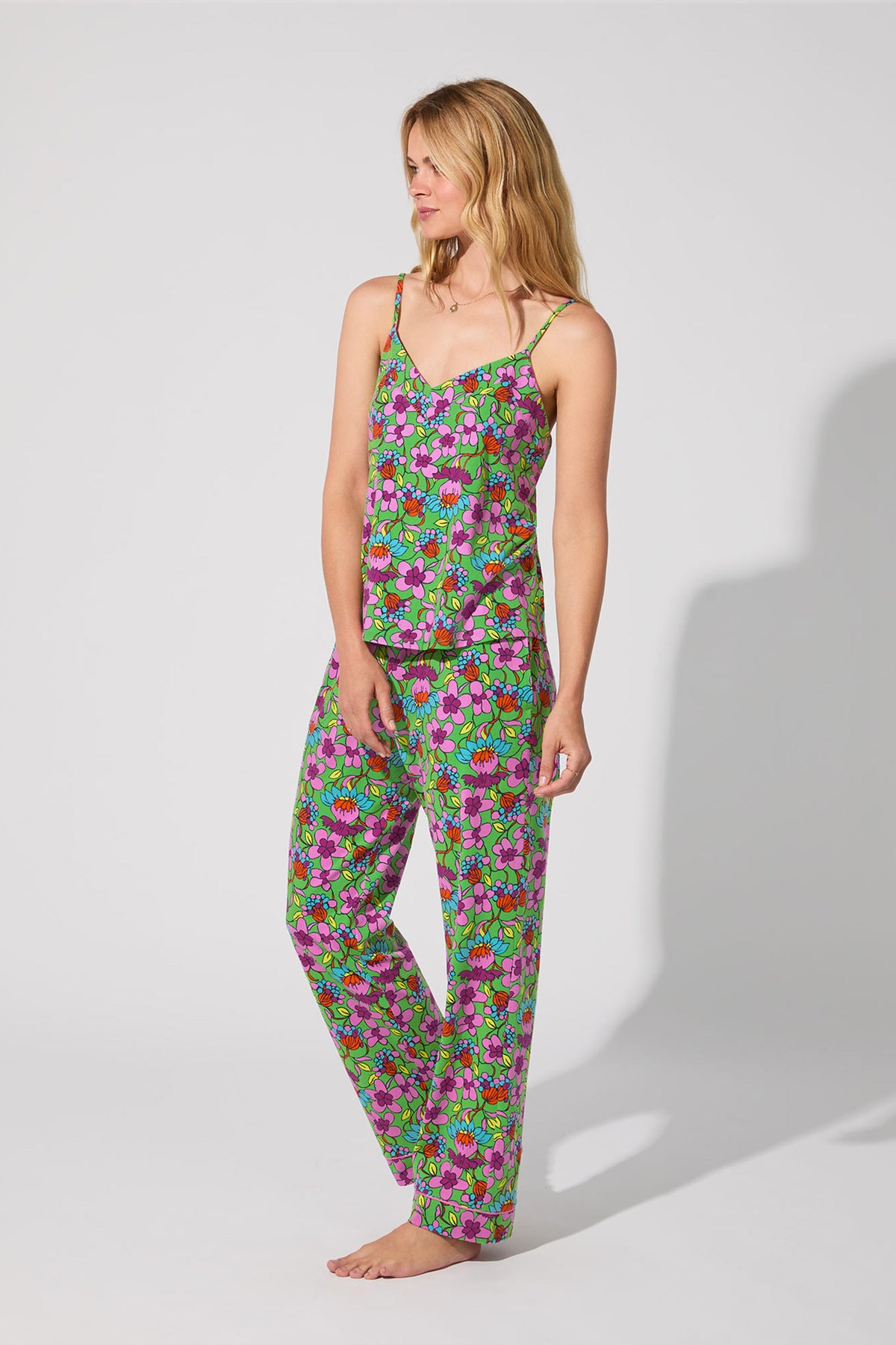 A woman stands barefoot against a plain background, wearing the Cami PJ Set - Garden Floral, which features a sleeveless camisole top and loose-fitting pants with colorful green, pink, purple, and orange floral patterns.