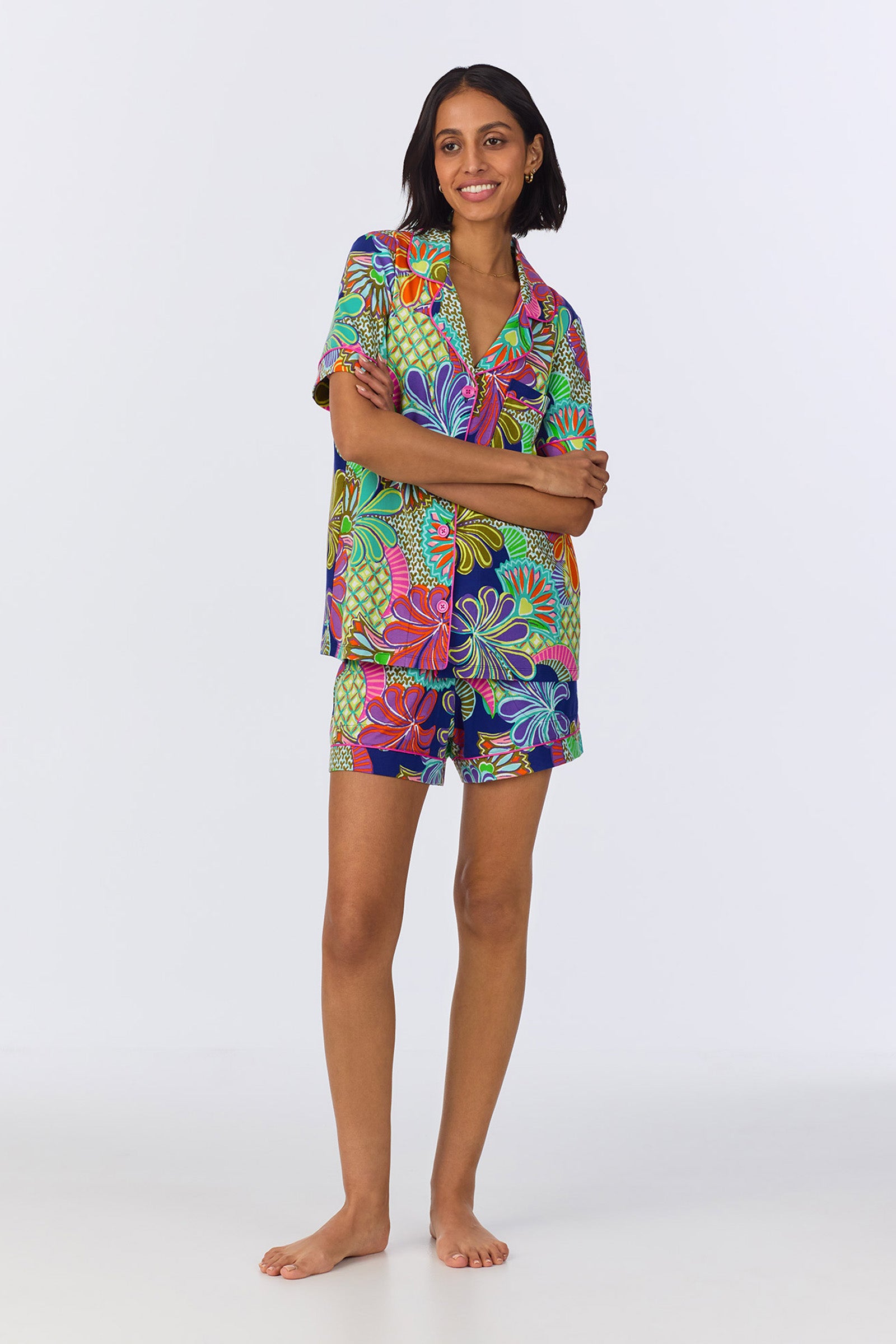 A woman stands barefoot, smiling with arms crossed, wearing the Shorty PJ Set - Zanzibar Stretch Jersey in Multi, a colorful short-sleeved pajama set featuring a vibrant floral pattern on a white background.