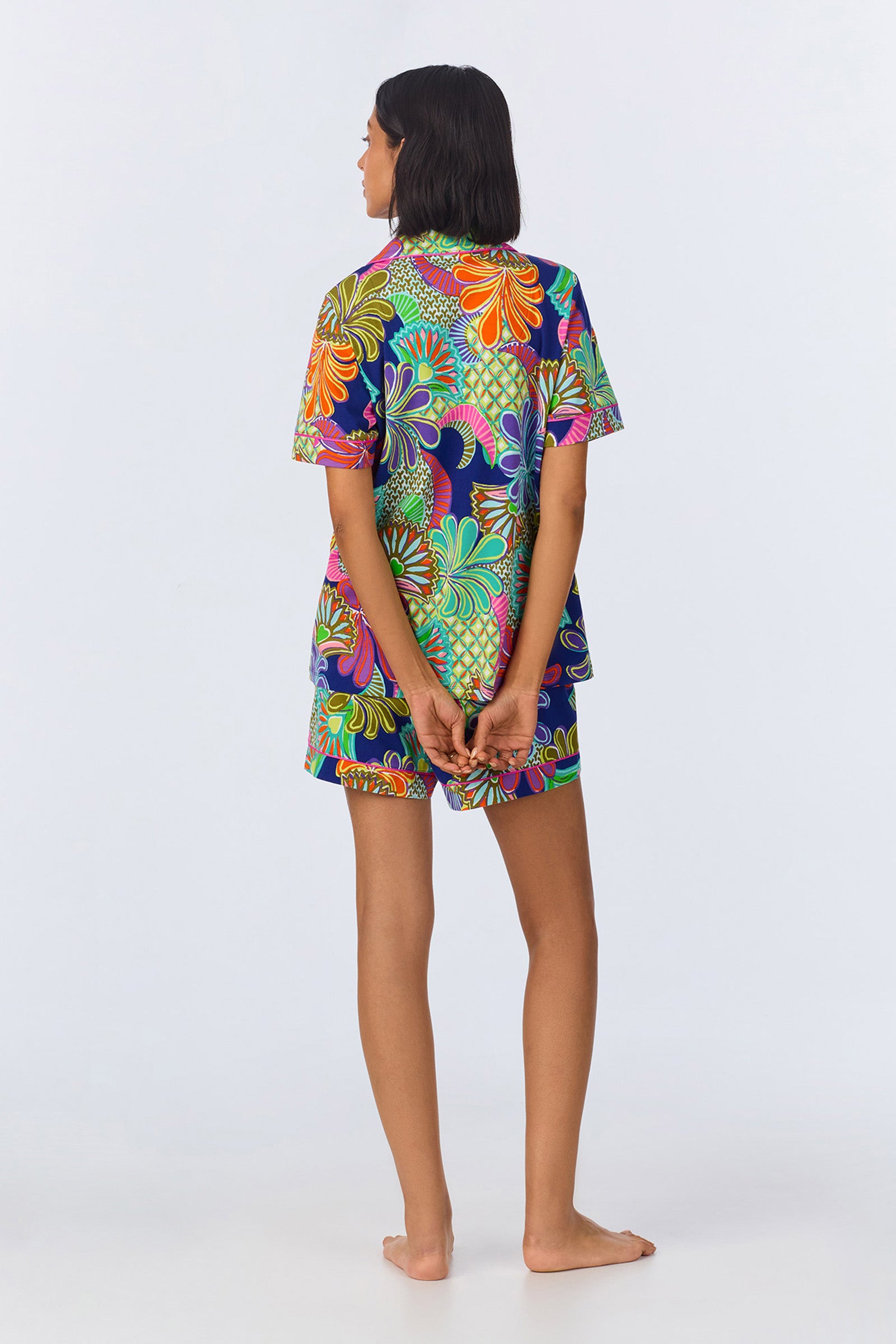 A person stands barefoot, facing away, with hands clasped behind their back. They wear the Shorty PJ Set - Zanzibar Stretch Jersey, featuring a colorful, bold floral pattern on a dark background.
