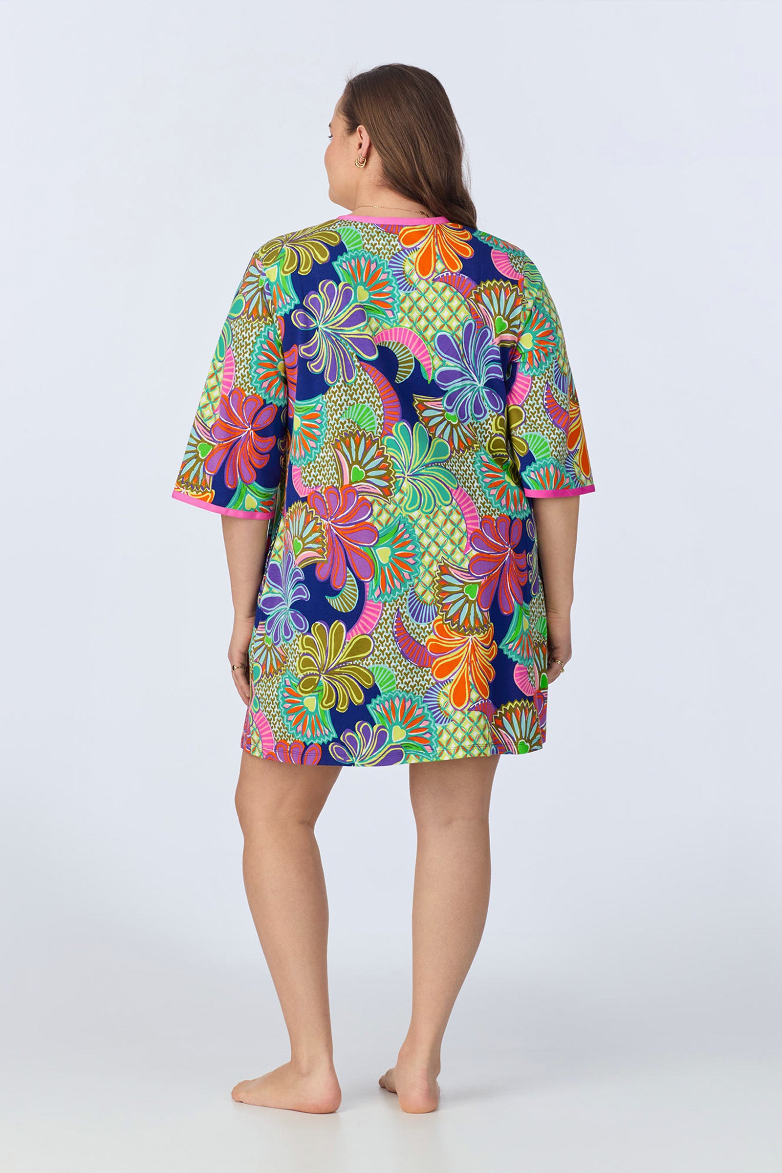 A woman stands barefoot, facing away, wearing the PJ Caftan Dress - Zanzibar Stretch Jersey, featuring a bold floral knee-length design in vibrant green, pink, orange, yellow, and purple on a dark background.