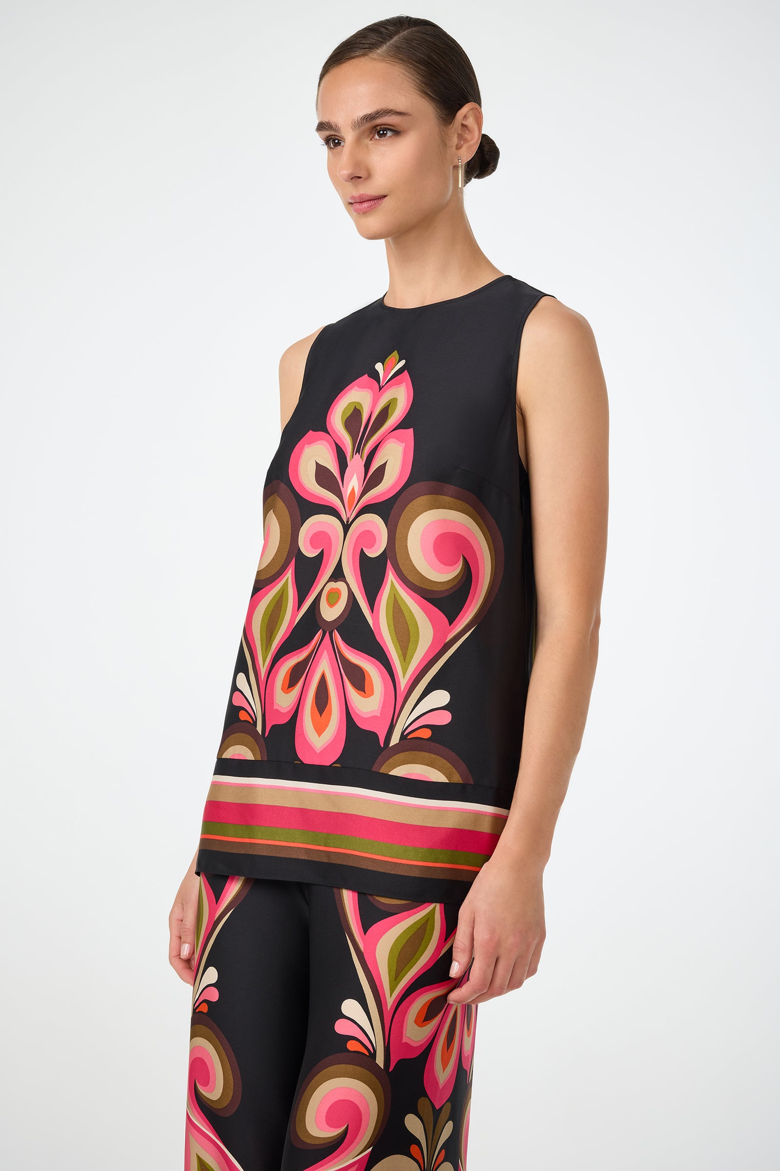 A woman with dark hair in a low bun wears the Malema Sleeveless Top in black, featuring a bold, colorful symmetrical floral pattern in pink, orange, green, and brown. She stands against a plain white background.