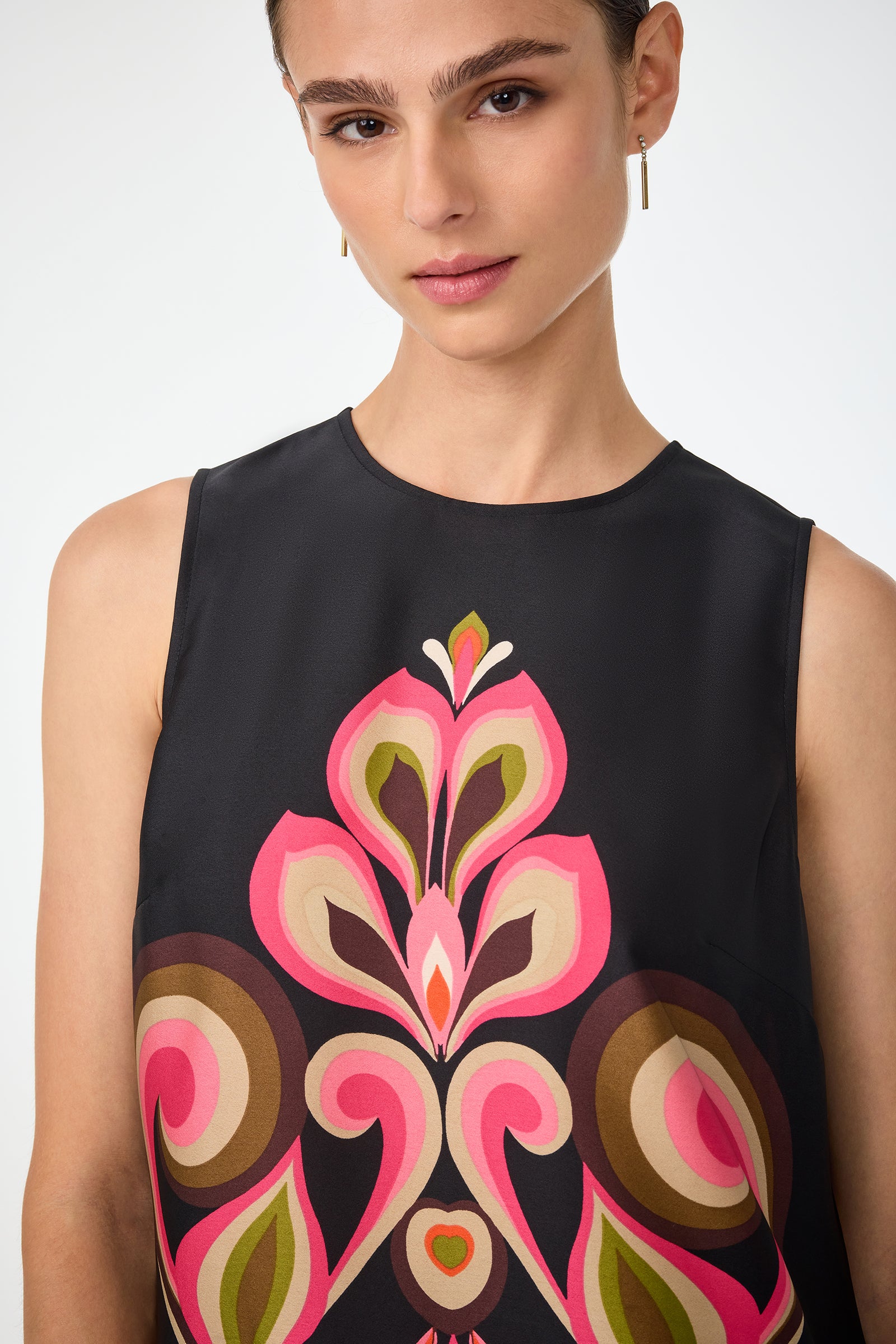 A woman wears the Malema Sleeveless Top, a black piece with a bold, abstract floral print in pink, brown, and green. She has light makeup, slicked-back hair, and gold rectangular drop earrings.