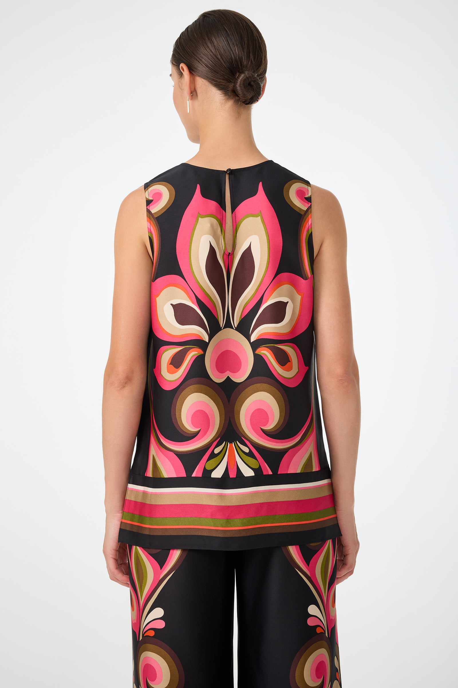 A woman with brown hair in a low bun is seen from the back, wearing the Malema Sleeveless Top and retro-patterned pants in pink, brown, green, and black. The background is plain white.