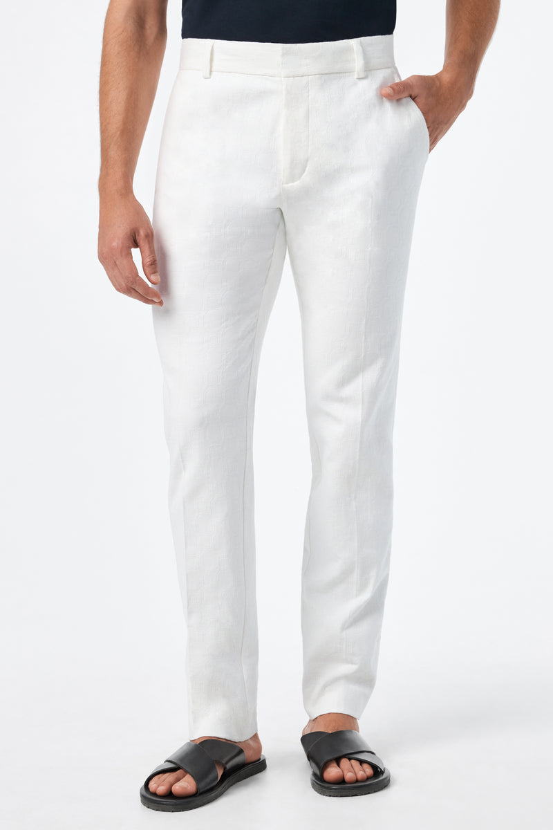 A person stands against a plain white background with one hand in their pocket, wearing the Clyde Slim Trouser - Stretch Herringbone, paired with a black shirt and black sandals.