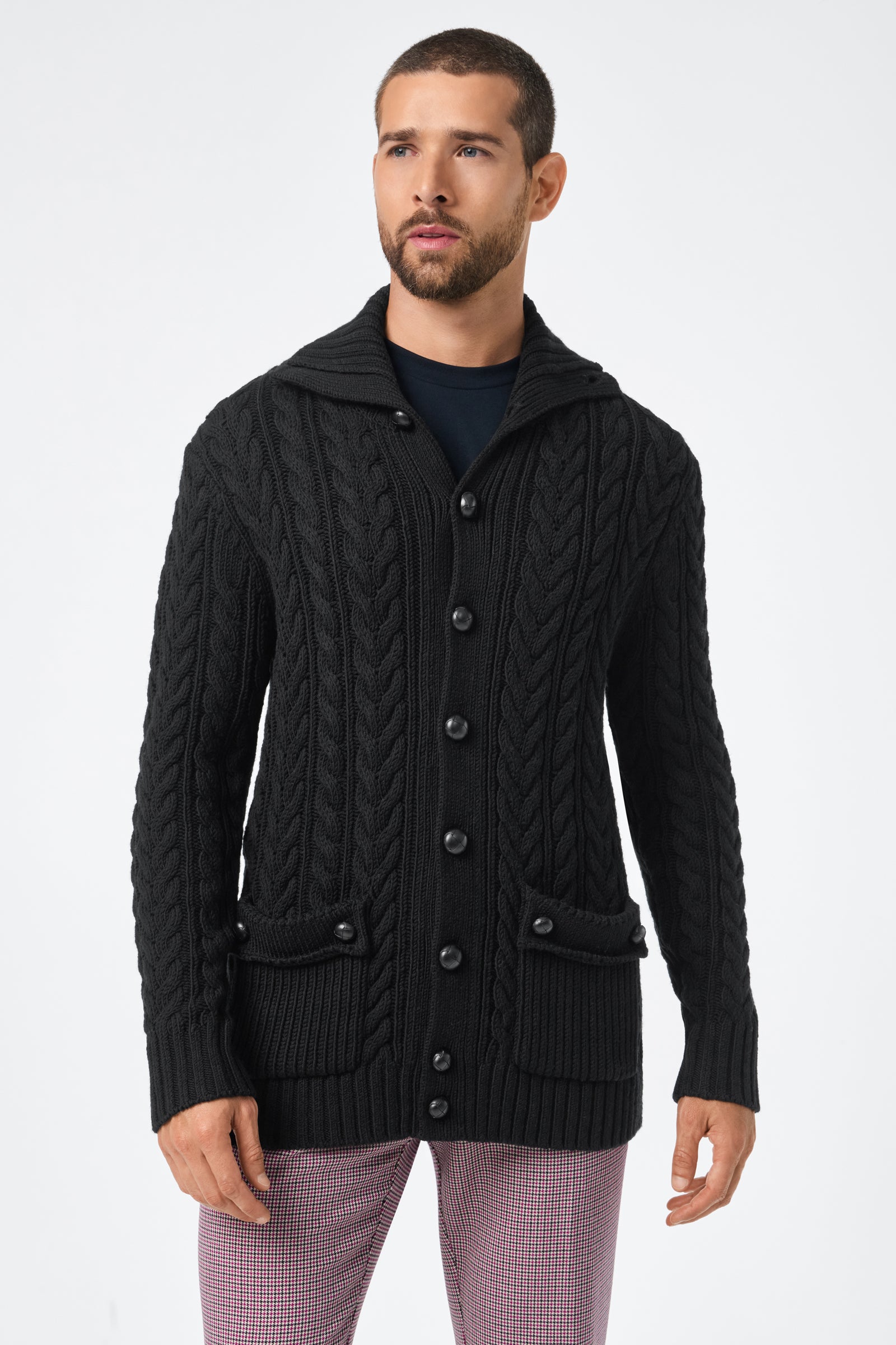 A man with short hair and a beard wears the black Raleigh Sweater Coat, featuring large buttons and front pockets, over a dark shirt with light plaid pants, standing against a plain white background.