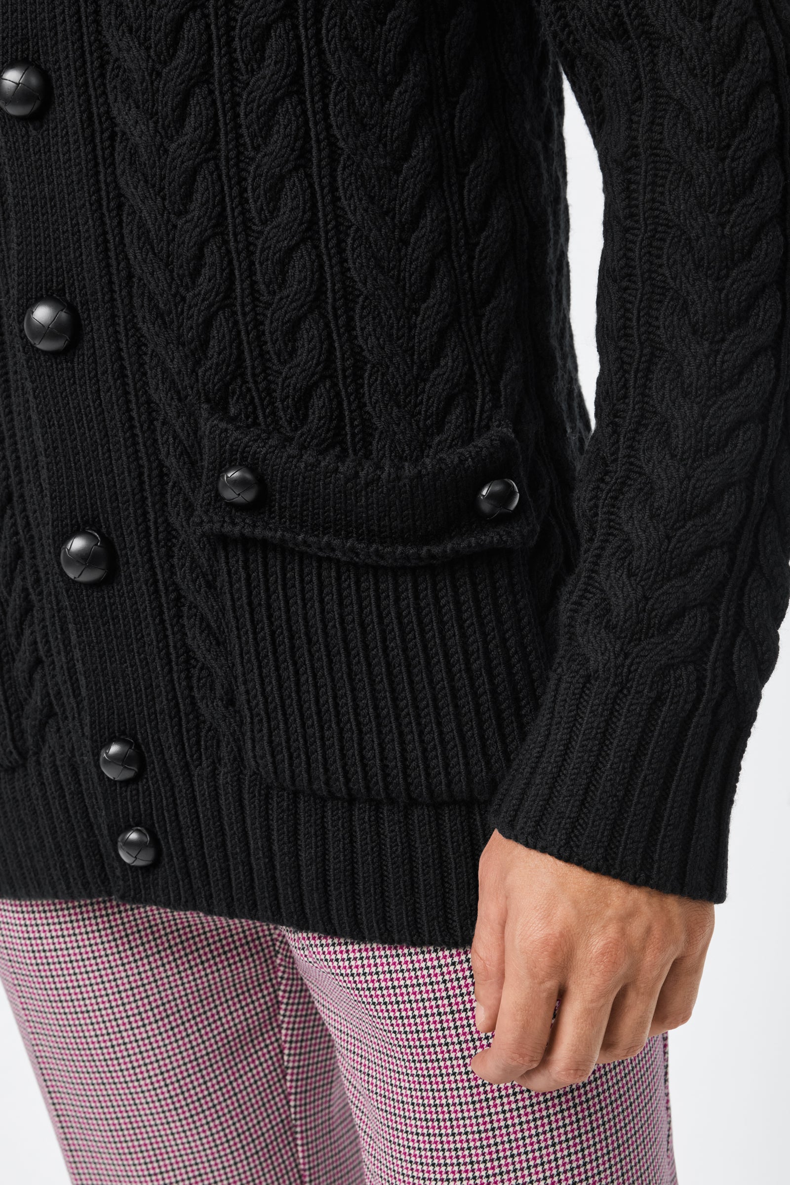 A person wears the Raleigh Sweater Coat, a black cable-knit cardigan with large black buttons and a pocket flap, paired with pink and white houndstooth pants. Only the torso and one hand are visible.