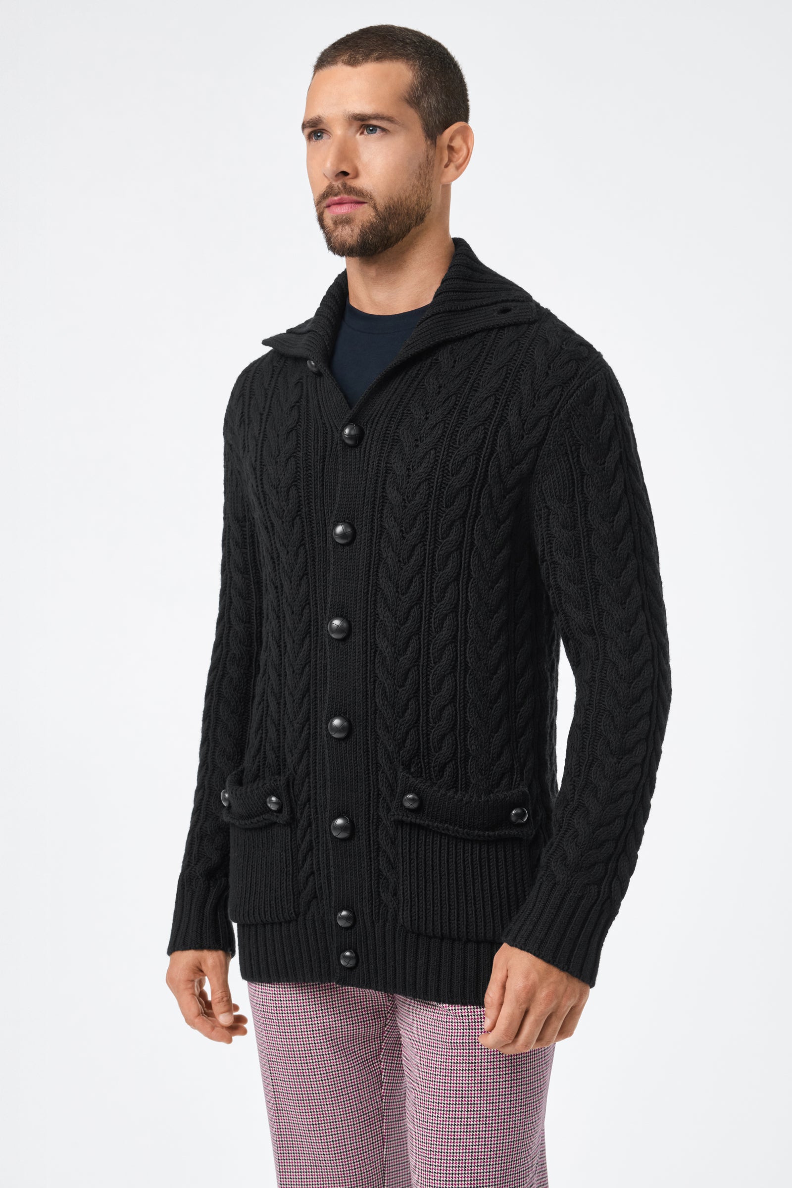 A man with short hair and a beard wears the Raleigh Sweater Coat—a black, chunky cable-knit button-up cardigan with front pockets—over a dark shirt and light plaid pants, standing against a plain white background.