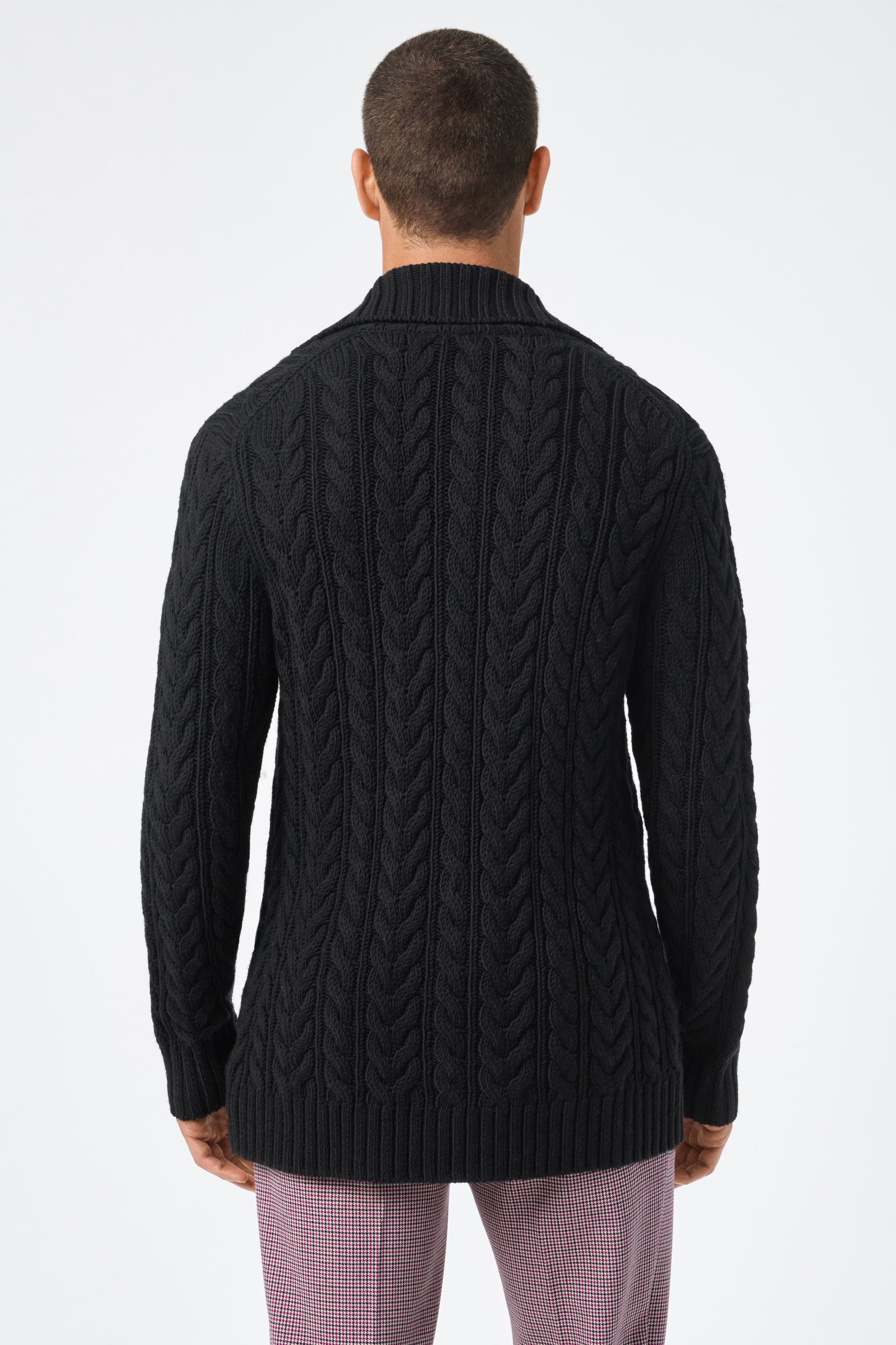 Someone is seen from behind wearing the Raleigh Sweater Coat, a black cable-knit sweater with ribbed hem and collar, paired with light pink plaid pants against a plain white background.