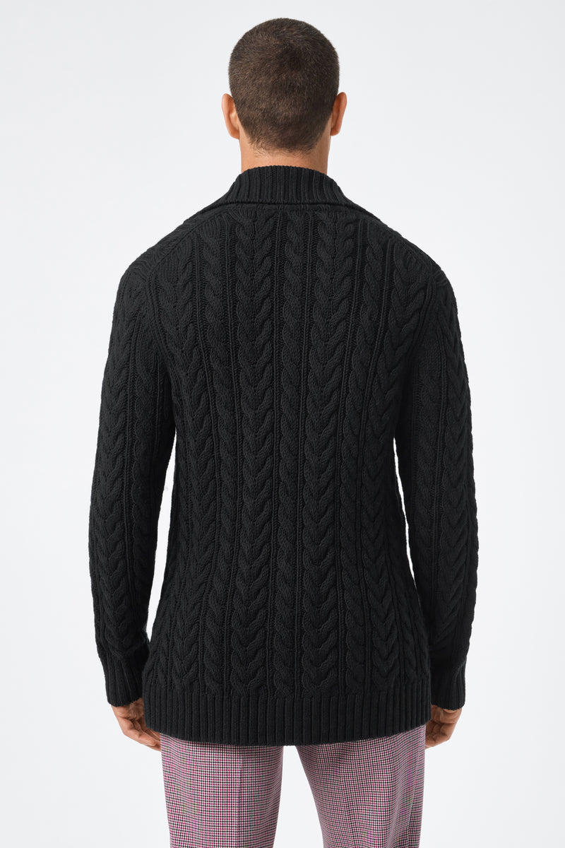 Someone is seen from behind wearing the Raleigh Sweater Coat, a black cable-knit sweater with ribbed hem and collar, paired with light pink plaid pants against a plain white background.