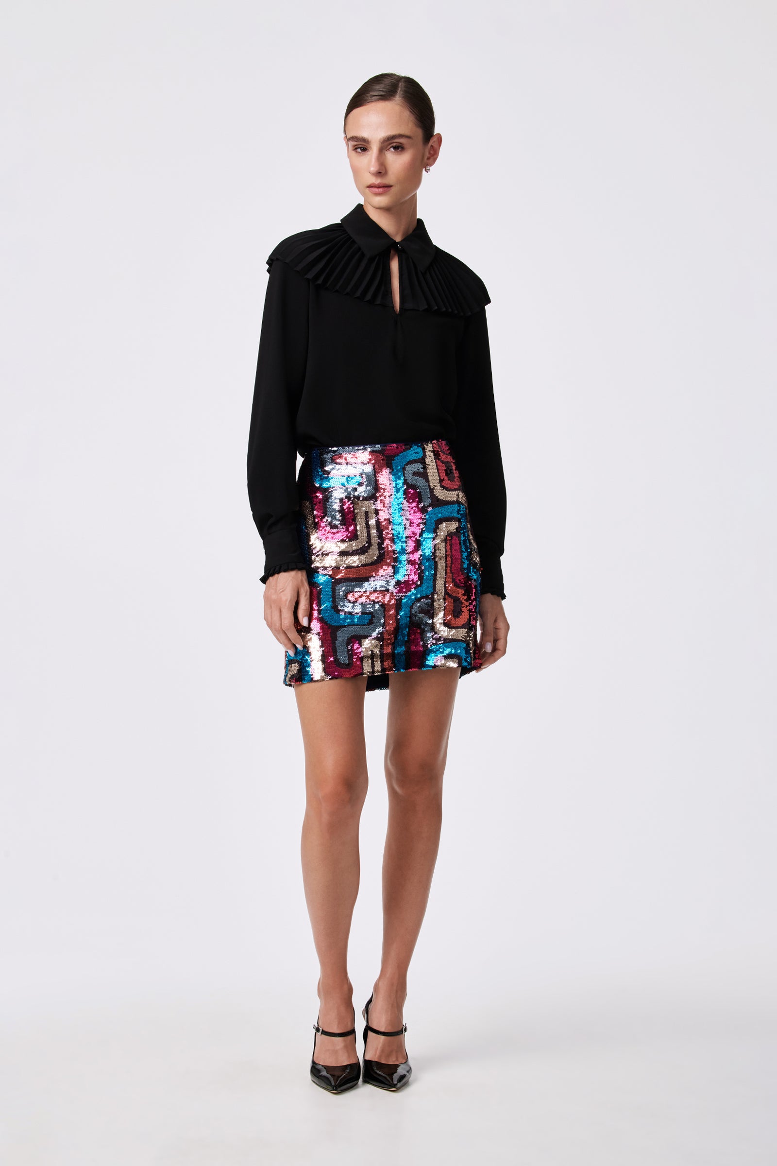 A woman stands against a plain background wearing the WINDMILL TOP, a black long-sleeve blouse with a pleated collar, paired with a shiny, multicolored sequined polyester skirt and black heels. Her hair is pulled back.