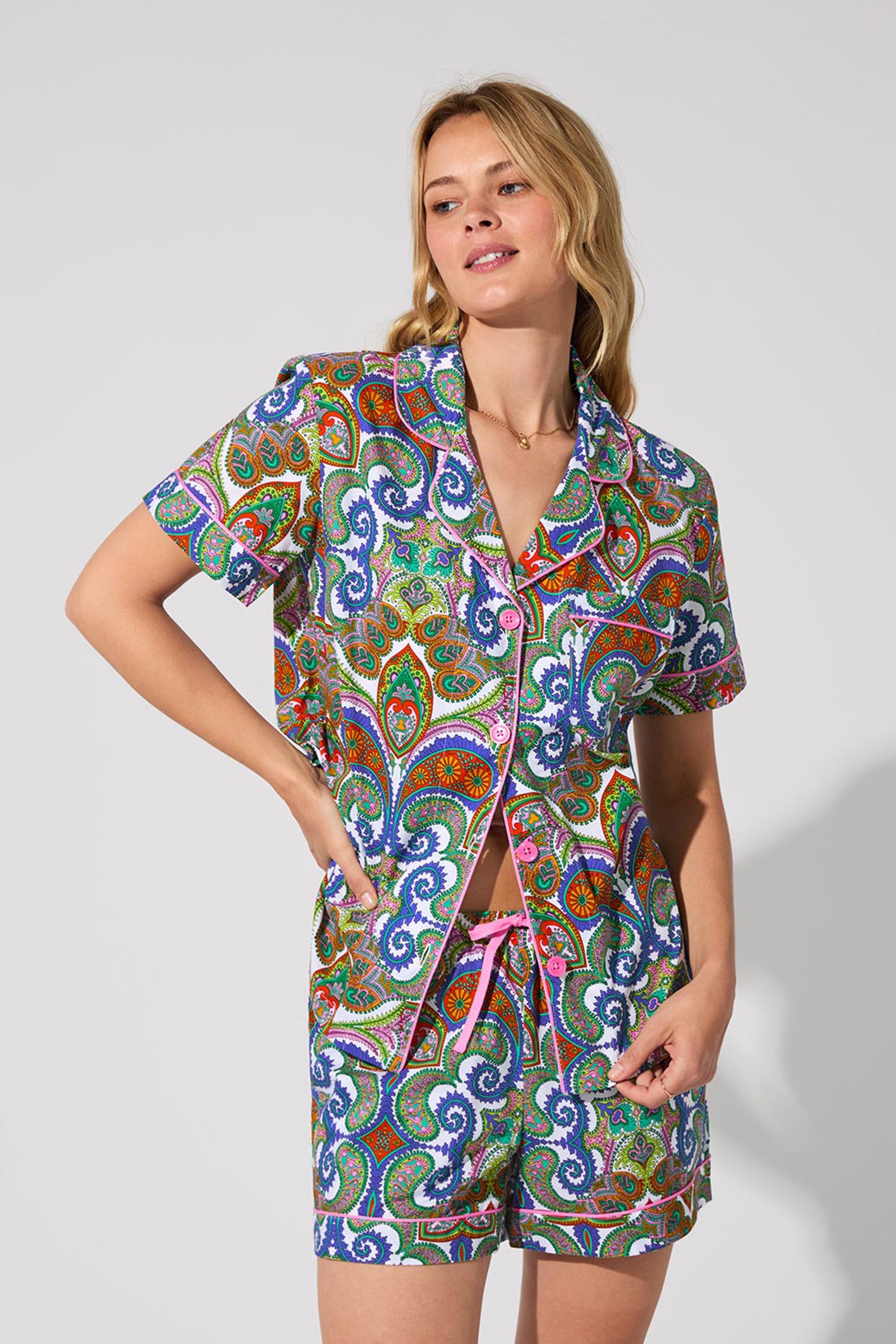 A woman with wavy blonde hair and a relaxed expression wears the Shorty PJ Set - Paisley, featuring a colorful paisley-patterned short-sleeve button-up shirt and matching shorts, standing against a plain light gray background.