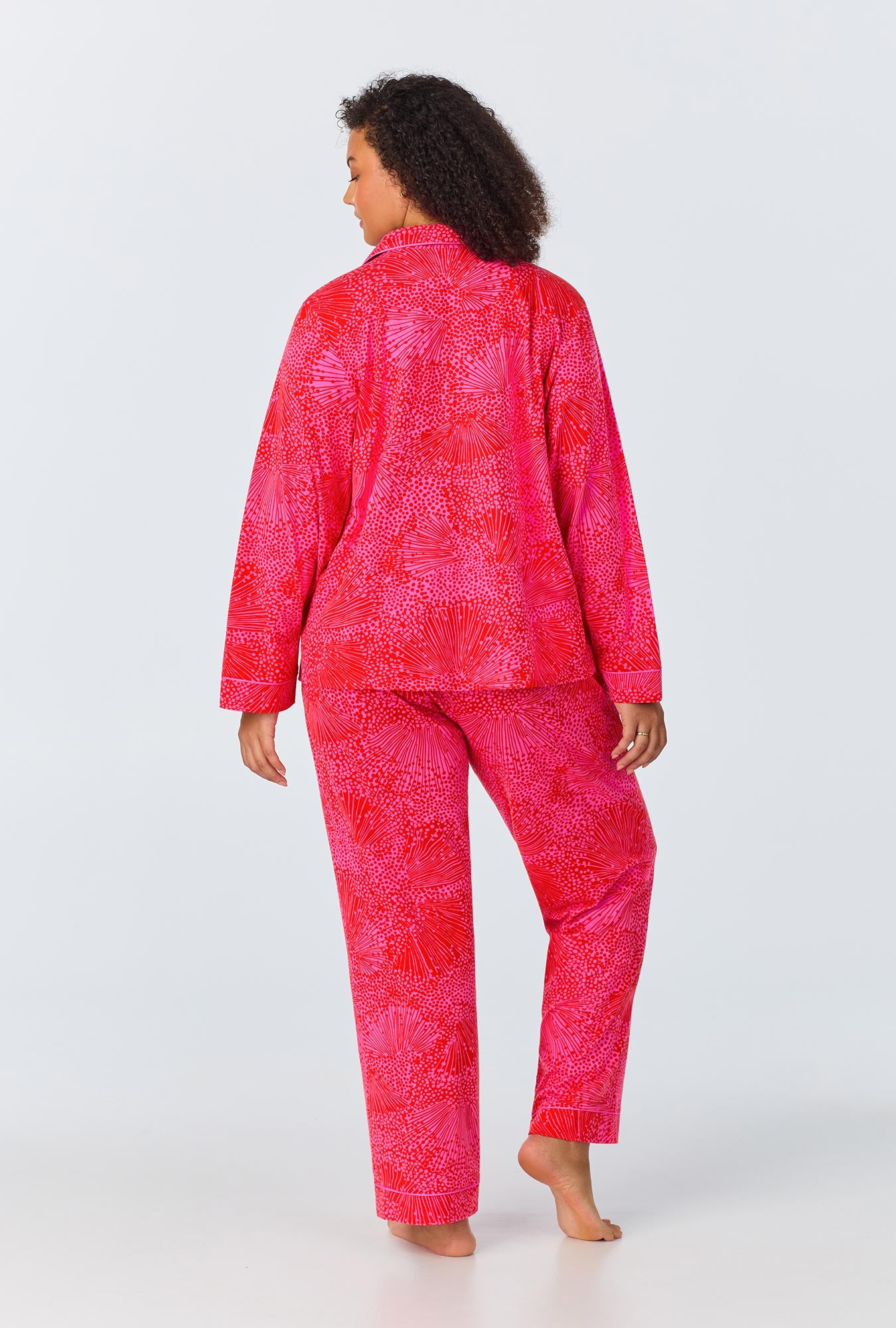 A woman with curly hair stands barefoot, facing away, wearing the Starburst Womens Long-Sleeve Long Pant Jersey PJ Set by Trina Turk in a bright pink and red pattern against a plain white background.
