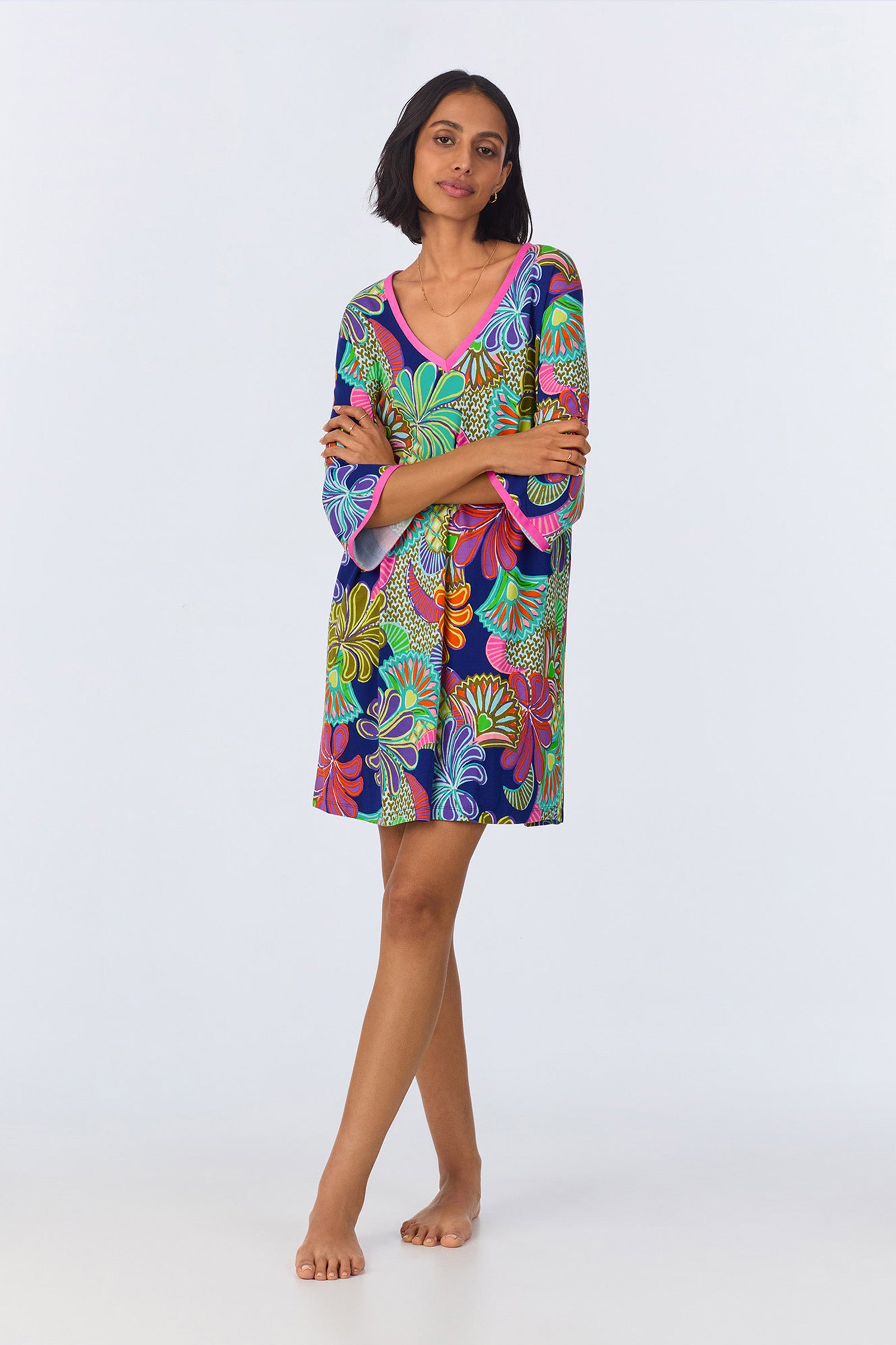 A woman with short dark hair stands barefoot against a plain background, arms crossed, wearing the PJ Caftan Dress - Zanzibar Stretch Jersey in Multi. The knee-length dress features vibrant abstract floral patterns.