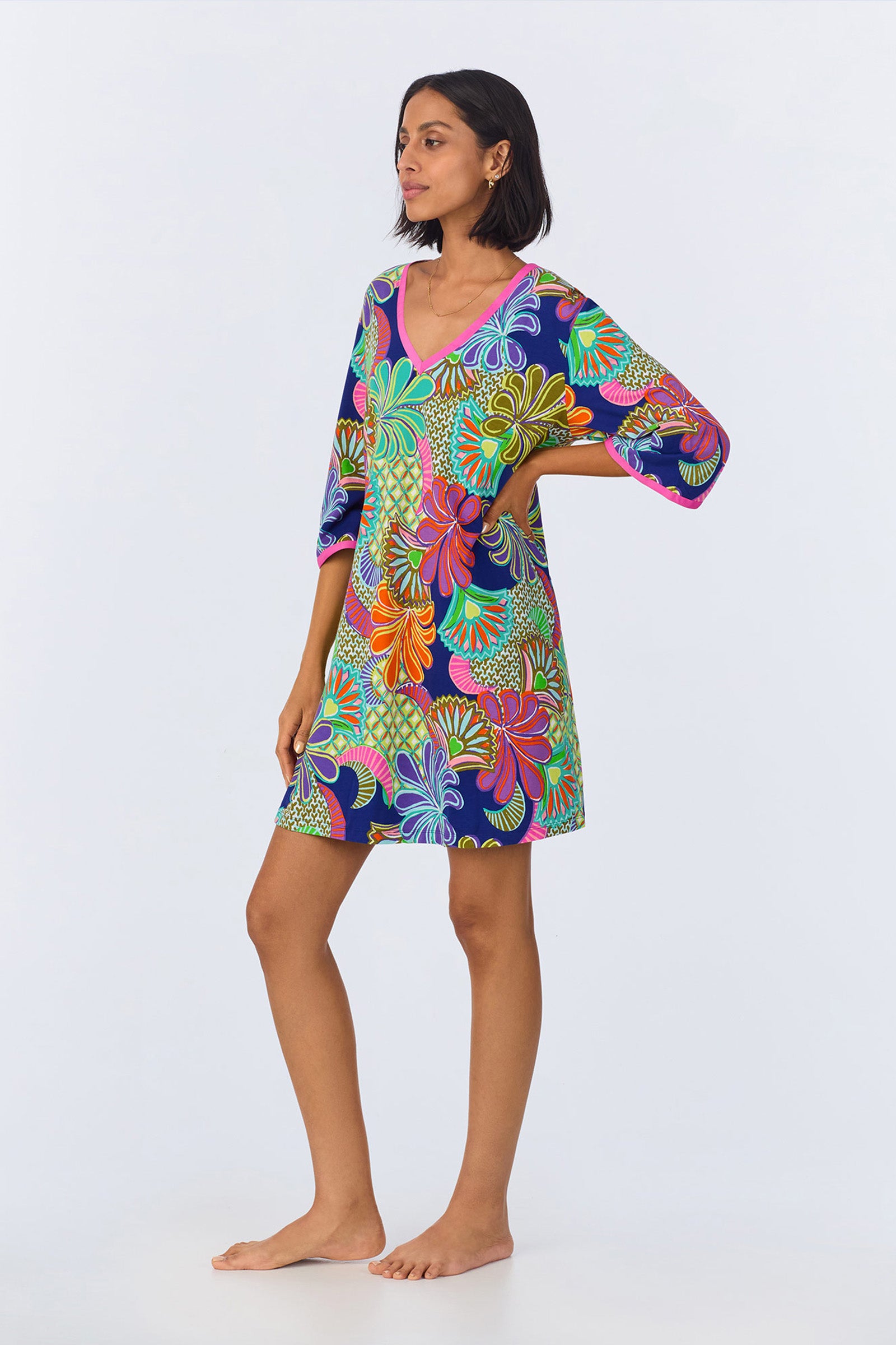 A woman stands barefoot, modeling the PJ Caftan Dress - Zanzibar Stretch Jersey, a knee-length dress with three-quarter sleeves, relaxed fit, and a bold abstract floral pattern in blue, green, orange, yellow, and purple.