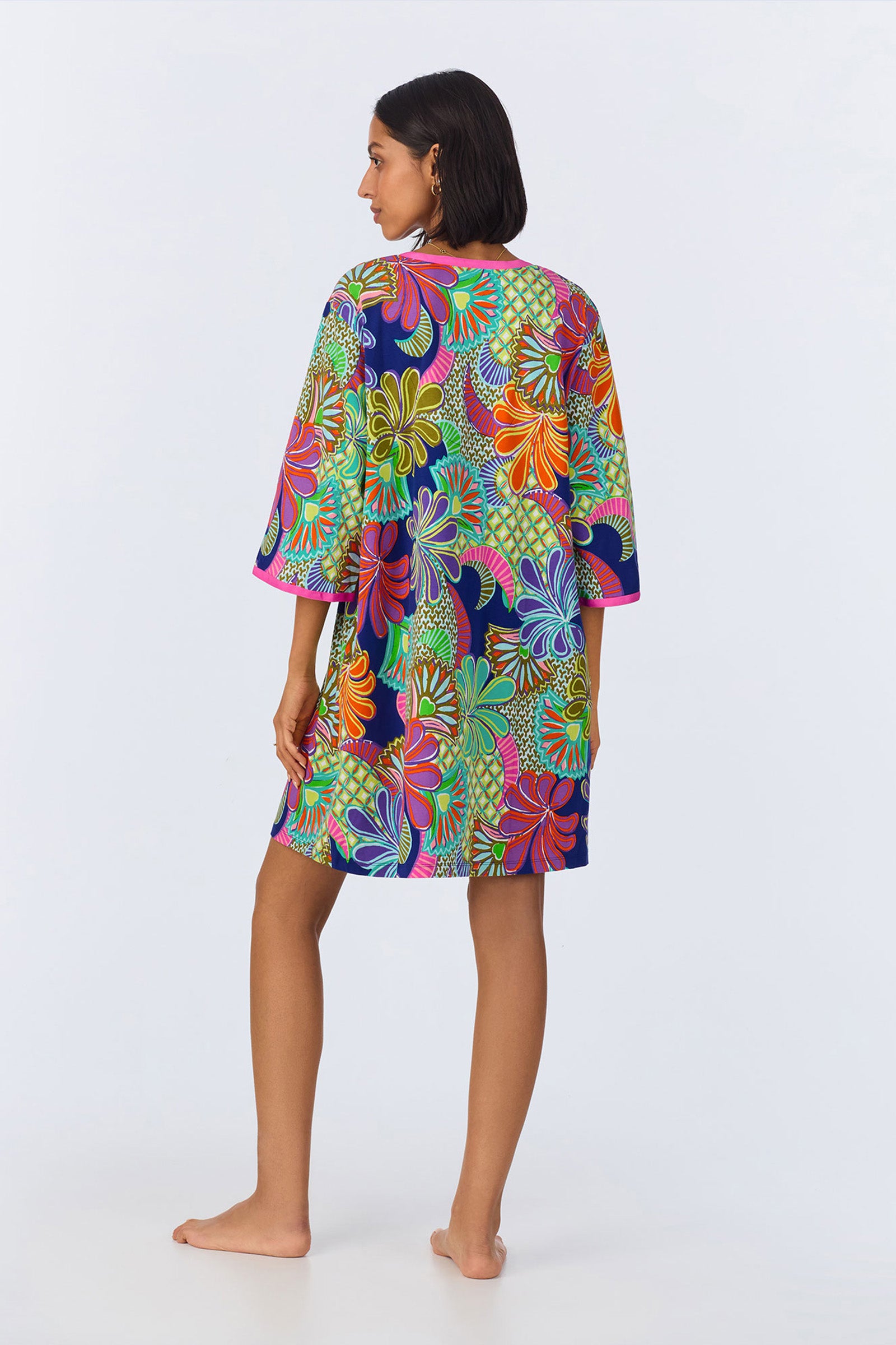 A woman stands barefoot, facing away, wearing the PJ Caftan Dress - Zanzibar Stretch Jersey, a colorful knee-length robe featuring a vibrant multicolored floral pattern against a plain light background.