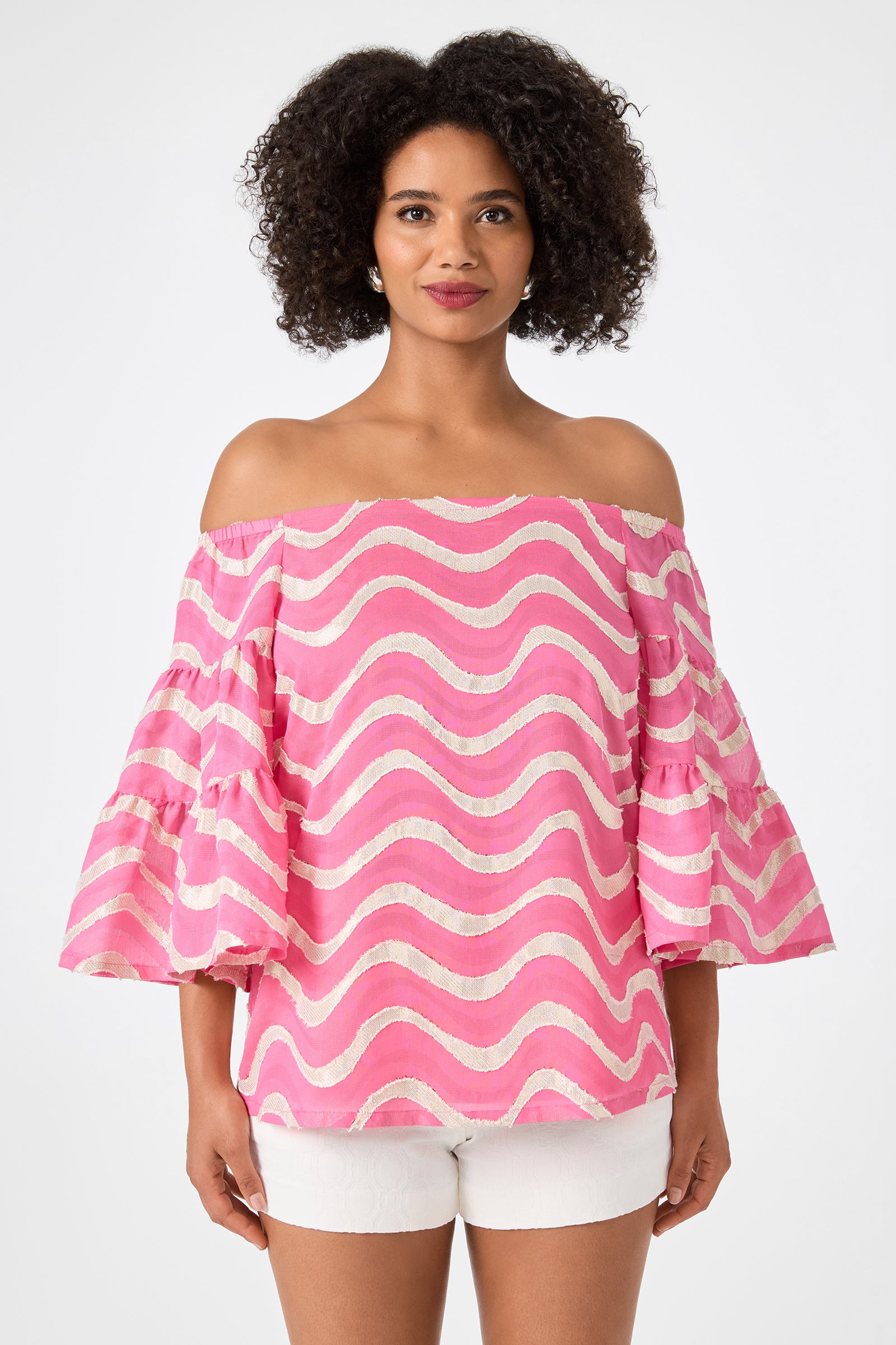 A woman models the Garnish 2 Off-Shoulder Top - Pinku, featuring pink and white wavy stripes and ruffled sleeves, paired with white shorts against a plain white background.
