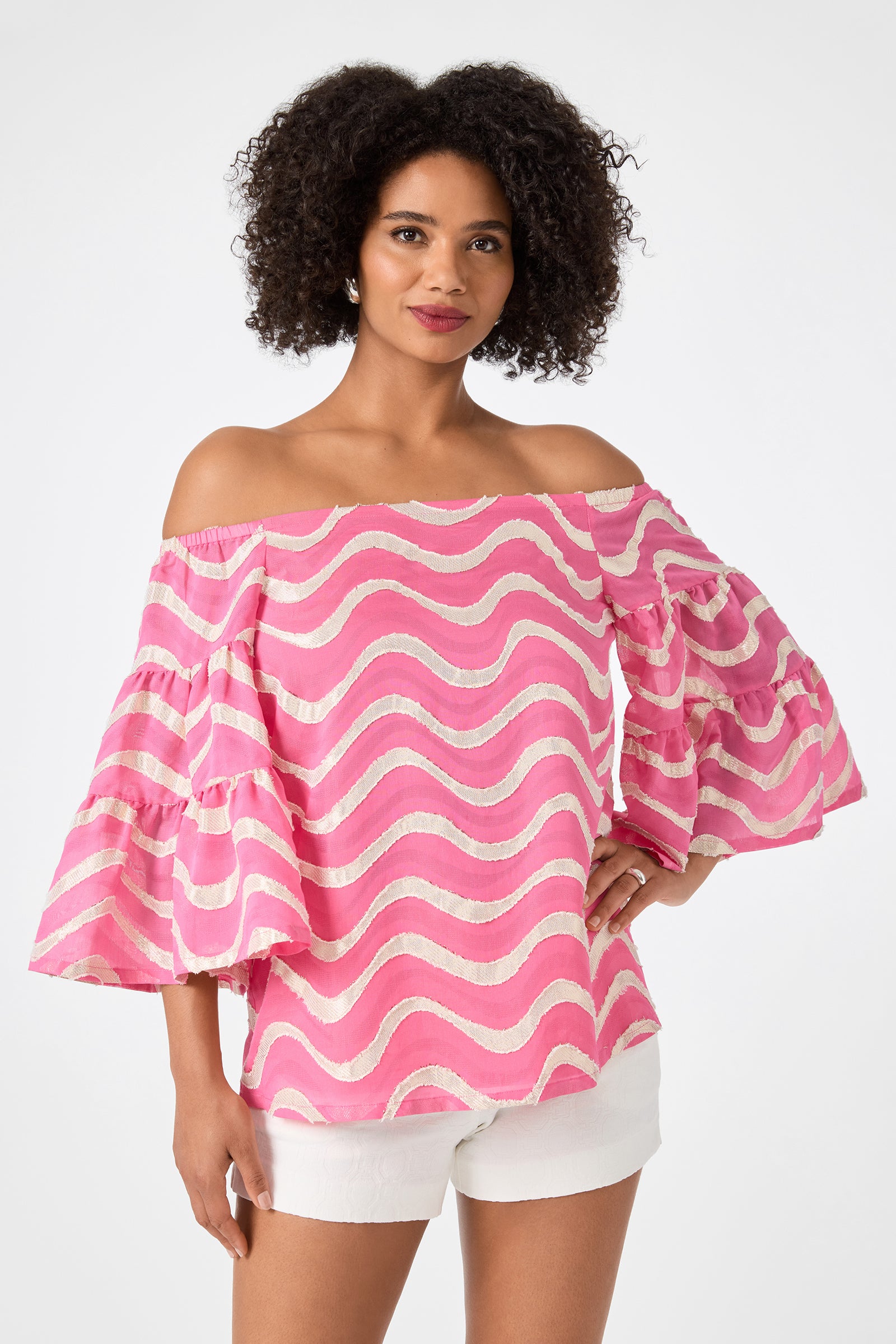 A woman smiles with one hand on her hip, wearing the Garnish 2 Off-Shoulder Top in Pinku with wavy stripes and ruffled sleeves, paired with white shorts against a plain white background.