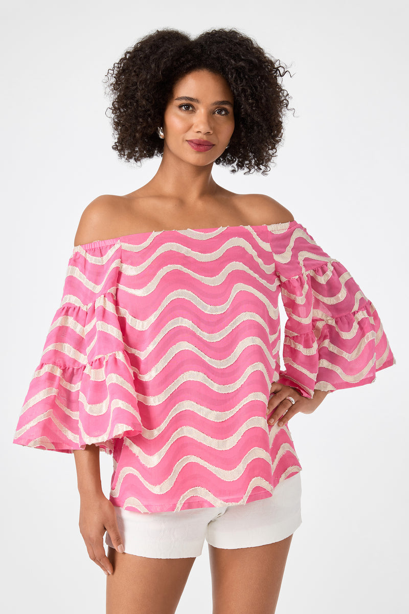 A woman smiles with one hand on her hip, wearing the Garnish 2 Off-Shoulder Top in Pinku with wavy stripes and ruffled sleeves, paired with white shorts against a plain white background.