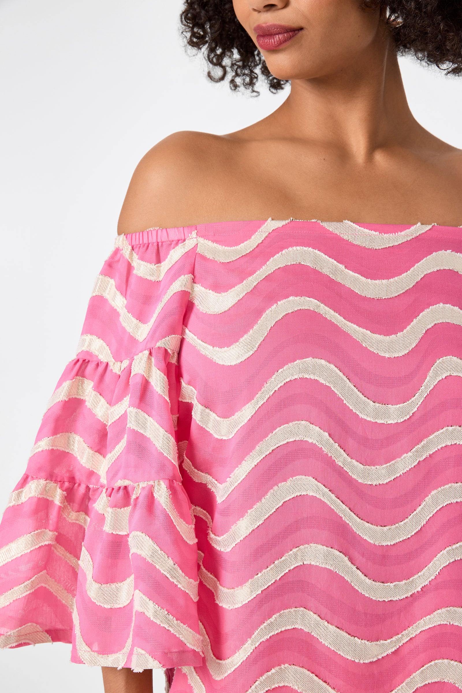 A woman models the Garnish 2 Off-Shoulder Top - Pinku, featuring white wavy stripes and ruffled sleeves, shown from shoulders to chest.