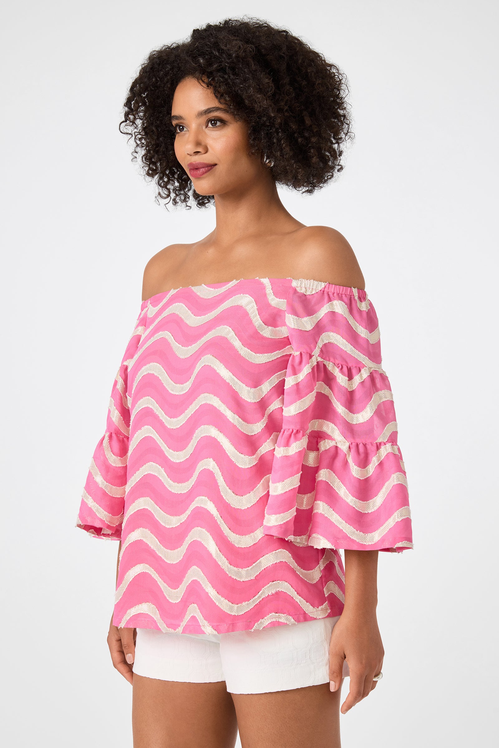 Model wears the Garnish 2 Off-Shoulder Top - Pinku, featuring pink and white wavy stripes and flared sleeves, paired with white shorts. She stands against a plain background looking to the side.