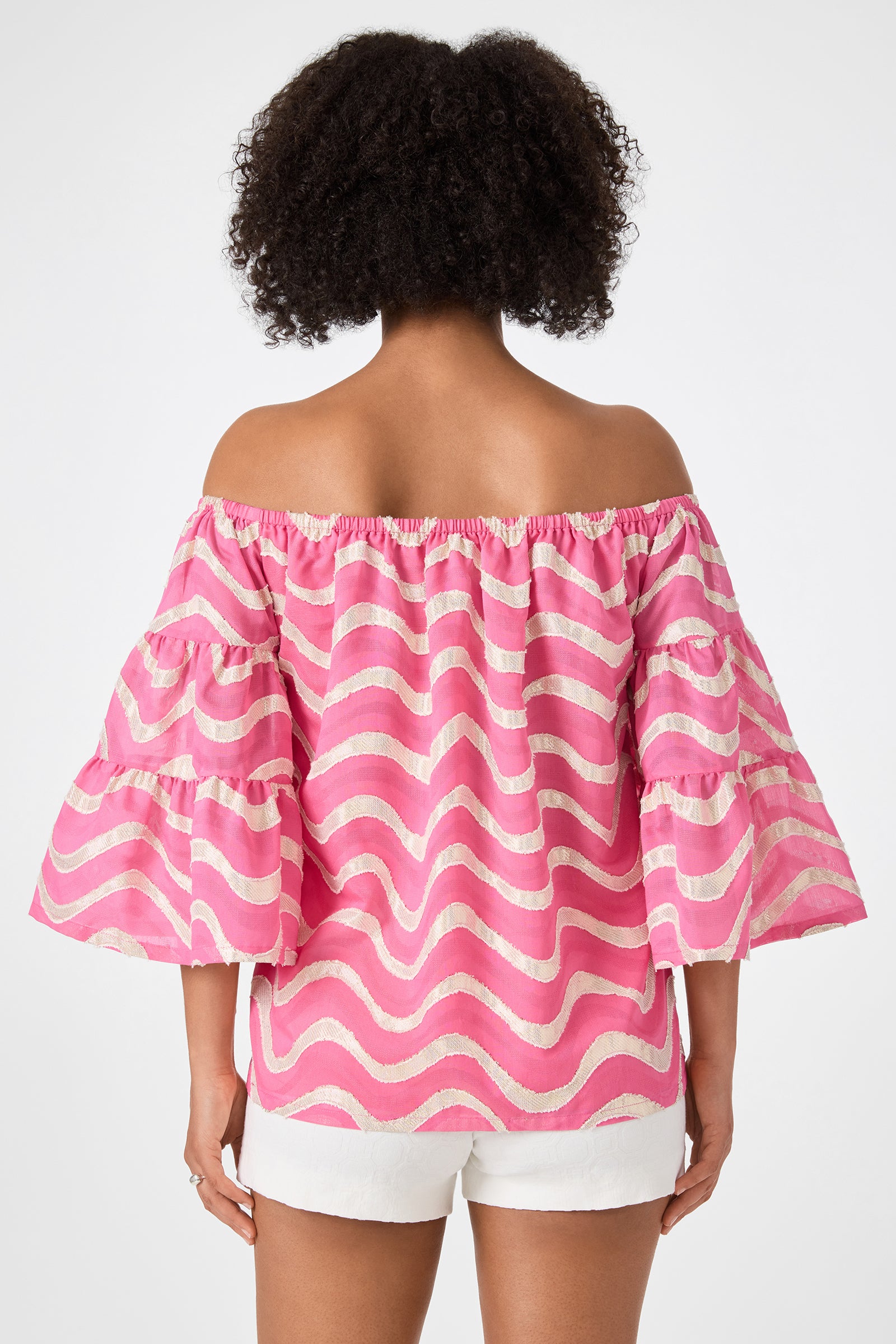 A person with curly hair is seen from the back wearing the Garnish 2 Off-Shoulder Top - Pinku, featuring white wavy stripes and ruffled sleeves, paired with white shorts against a plain background.