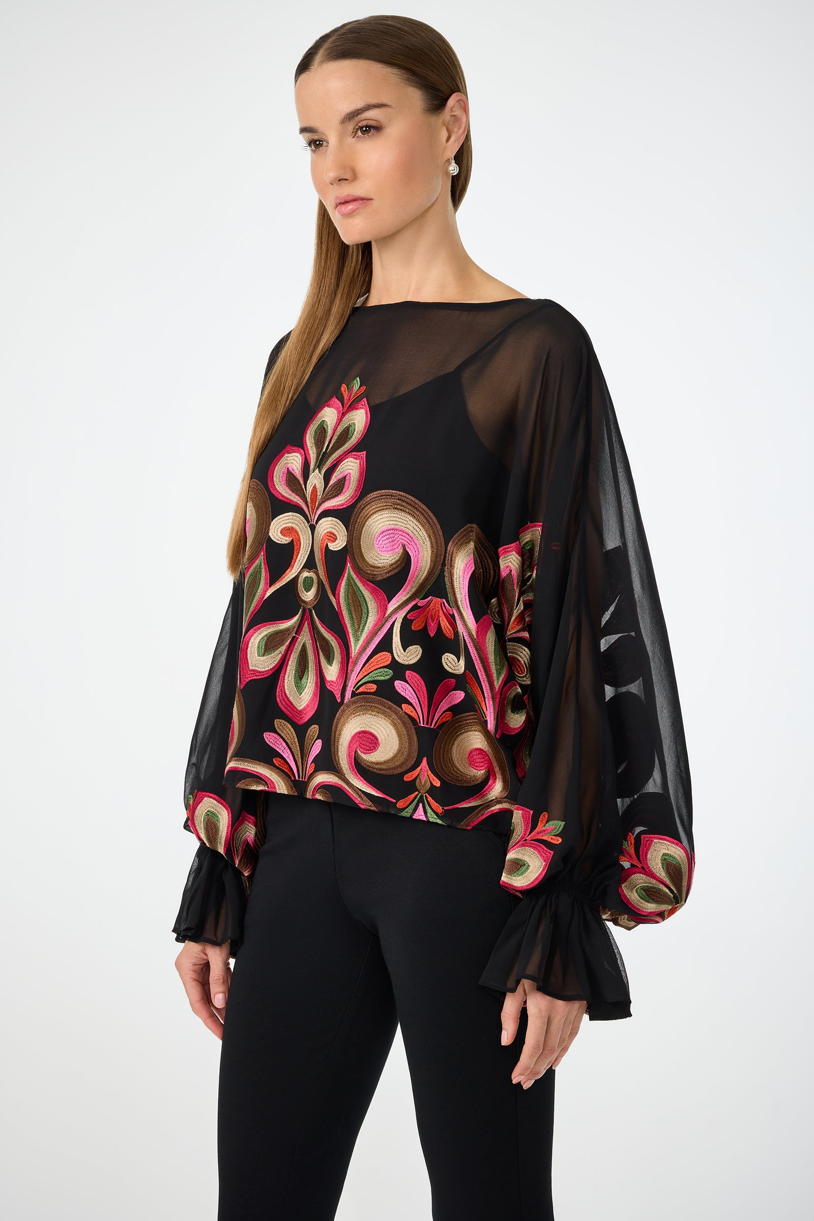 A woman with long, straight hair wears the Soho Embroidered Top by Sanctuary—a sheer black blouse with colorful embroidery and ruffled cuffs—paired with black pants against a plain light background.
