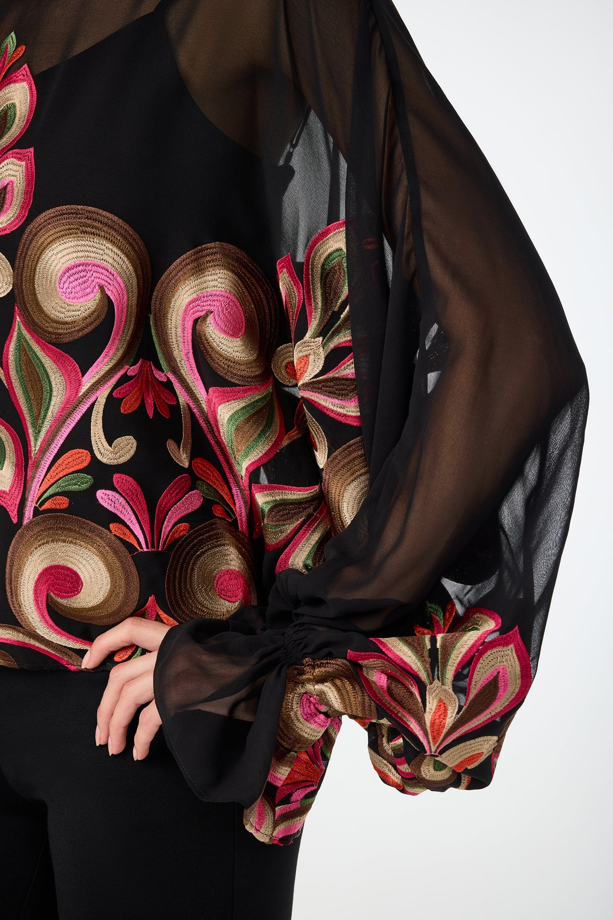 Wearing the Soho Embroidered Top by Sanctuary, a sheer black blouse with pink, green, and gold floral embroidery and voluminous sleeves, the person rests a hand on their hip.