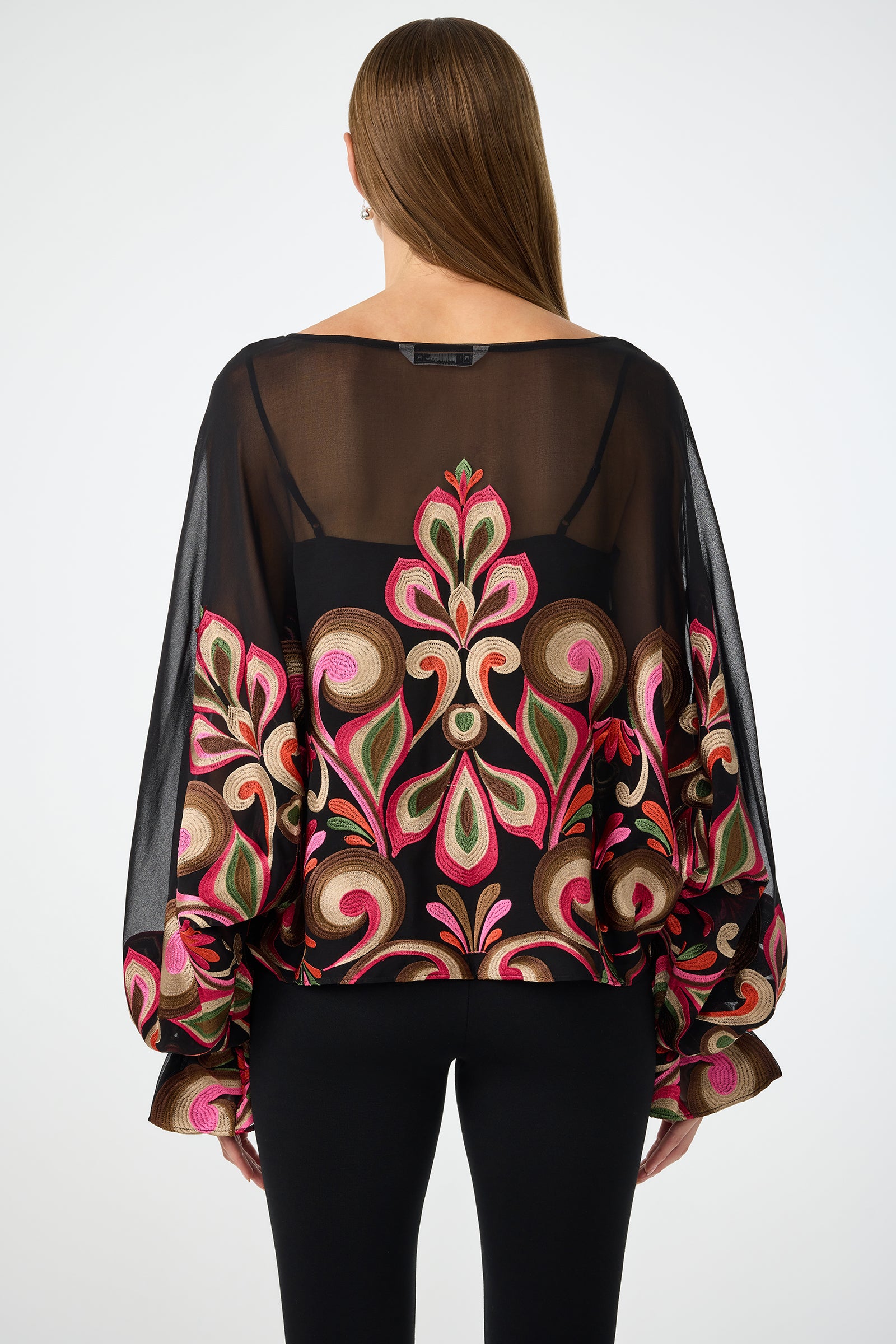 A woman with straight brown hair faces away from the camera, showcasing the back of the Sanctuary Soho Embroidered Top—a sheer black blouse with colorful floral embroidery and billowy sleeves—paired with black pants.