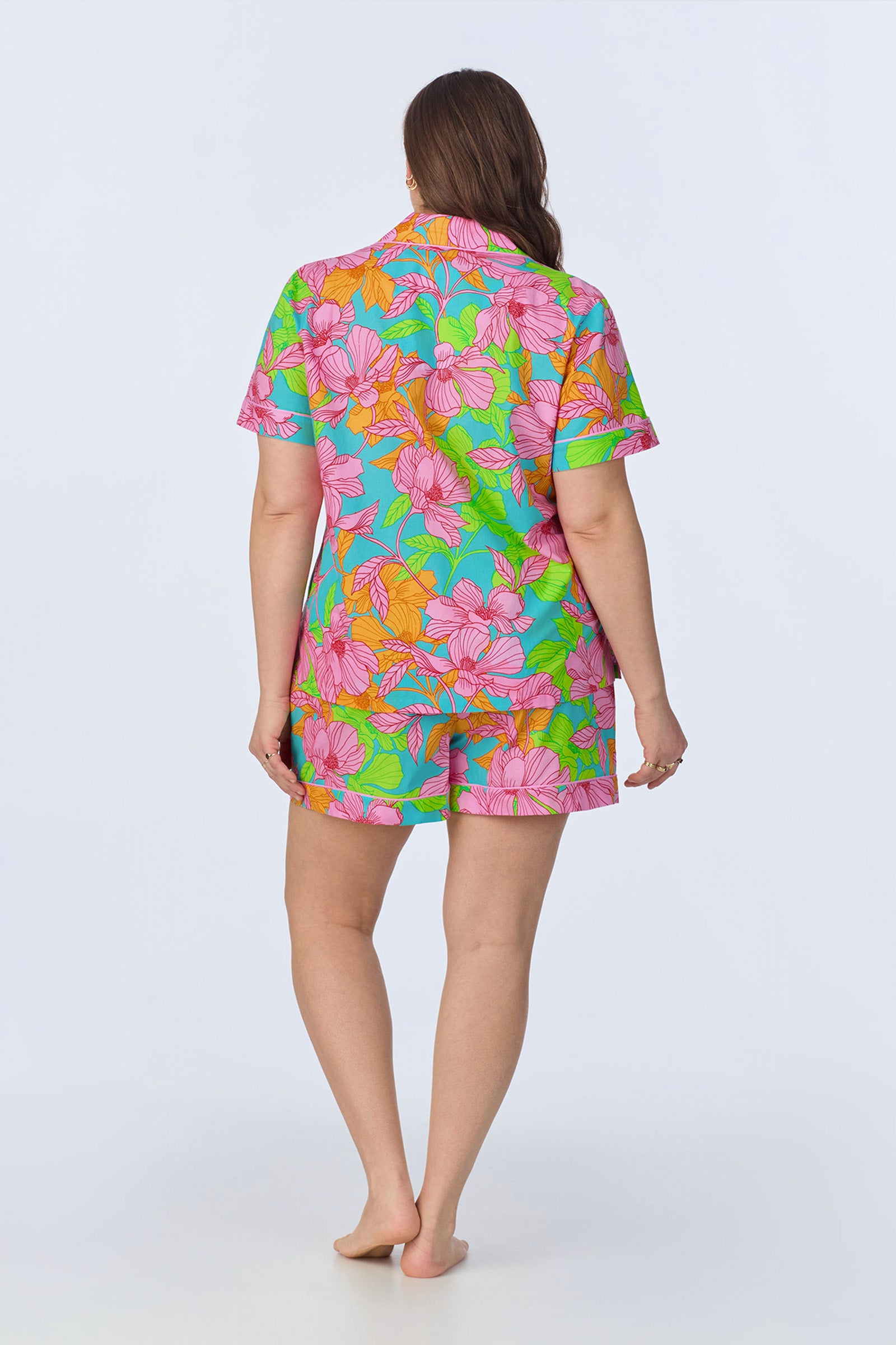 A woman stands barefoot, facing away, wearing the Shorty PJ Set - Linear Floral Poplin, a colorful matching short-sleeve shirt and shorts with a pink, orange, and green floral pattern on a blue background.