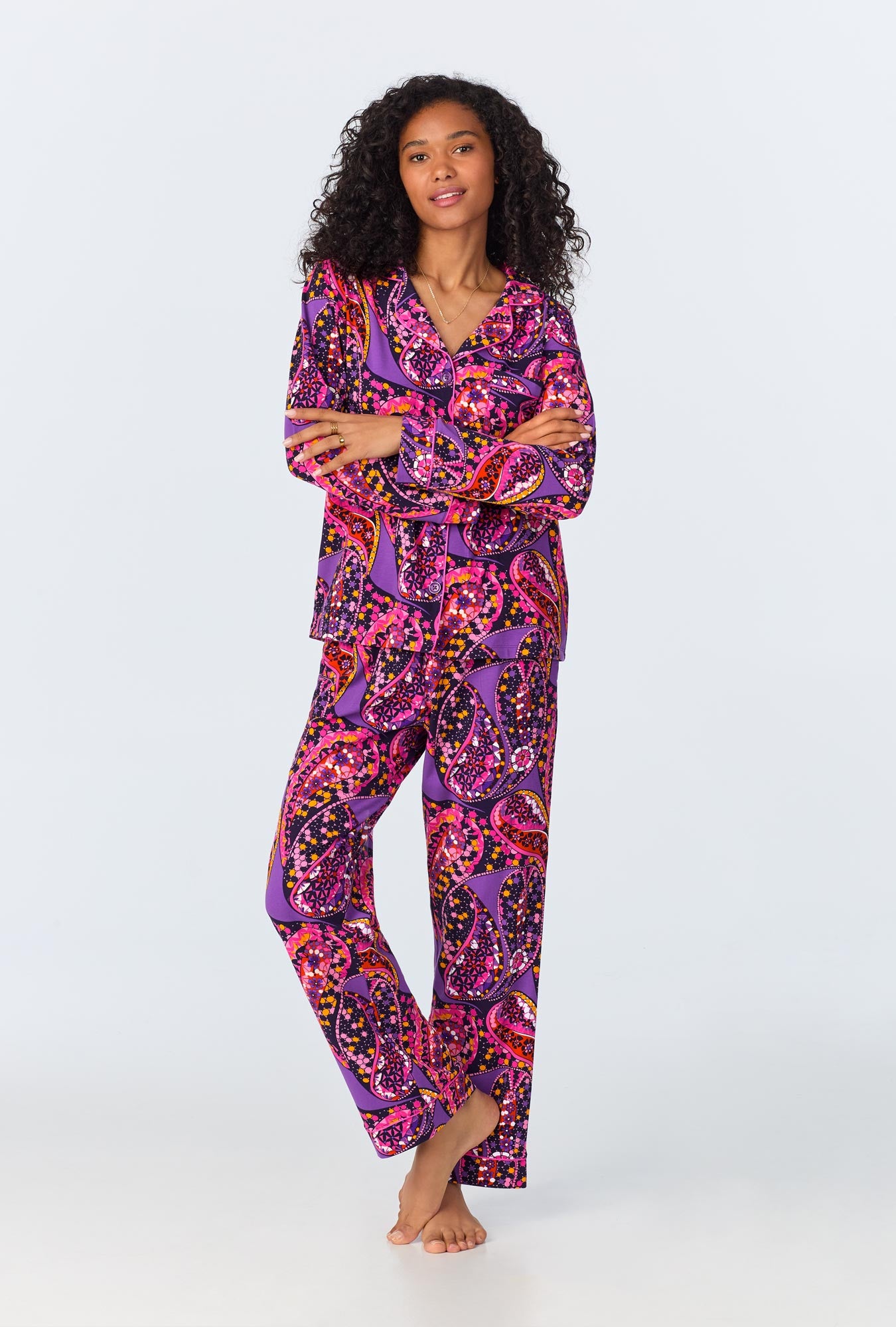 A woman with curly hair stands barefoot in the Tangled Paisley Womens Long Sleeve Long Pant Jersey PJ Set in Multi, featuring vibrant colorful patterns, against a plain light background.